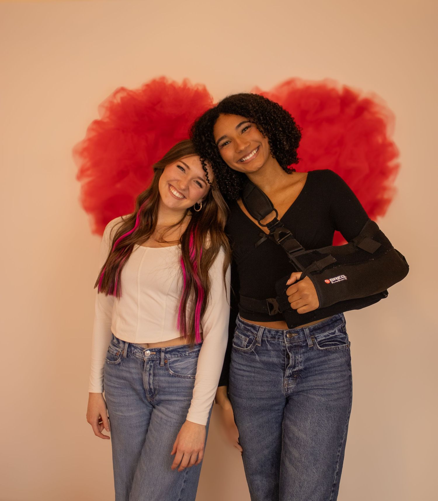 Two friends in matching jeans and contrasting tops pose playfully with bright red hair accessories against a beige wall.