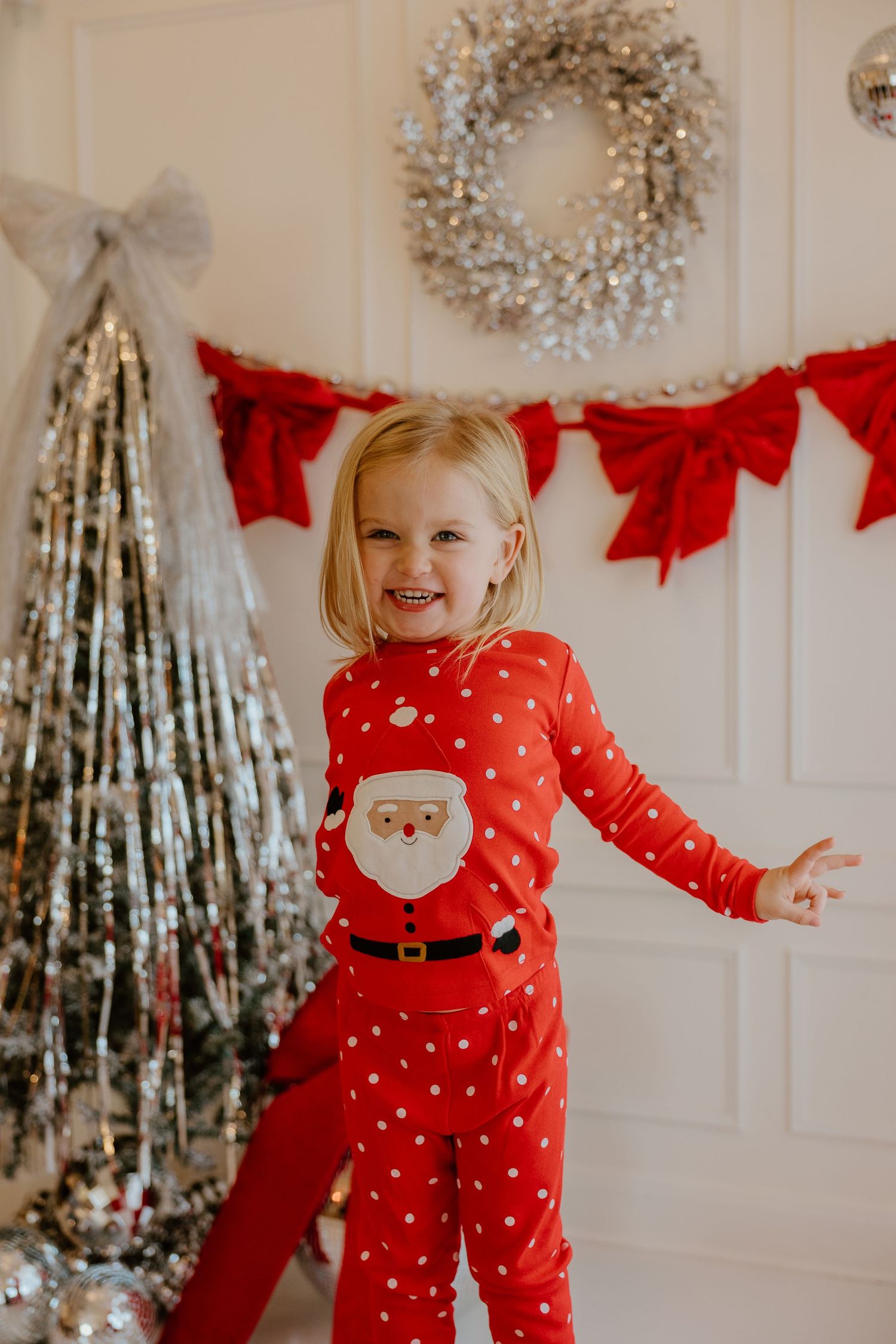 Red polka dot Santa pajamas shown in front of a silver Christmas tree and festive holiday decor with red bows.