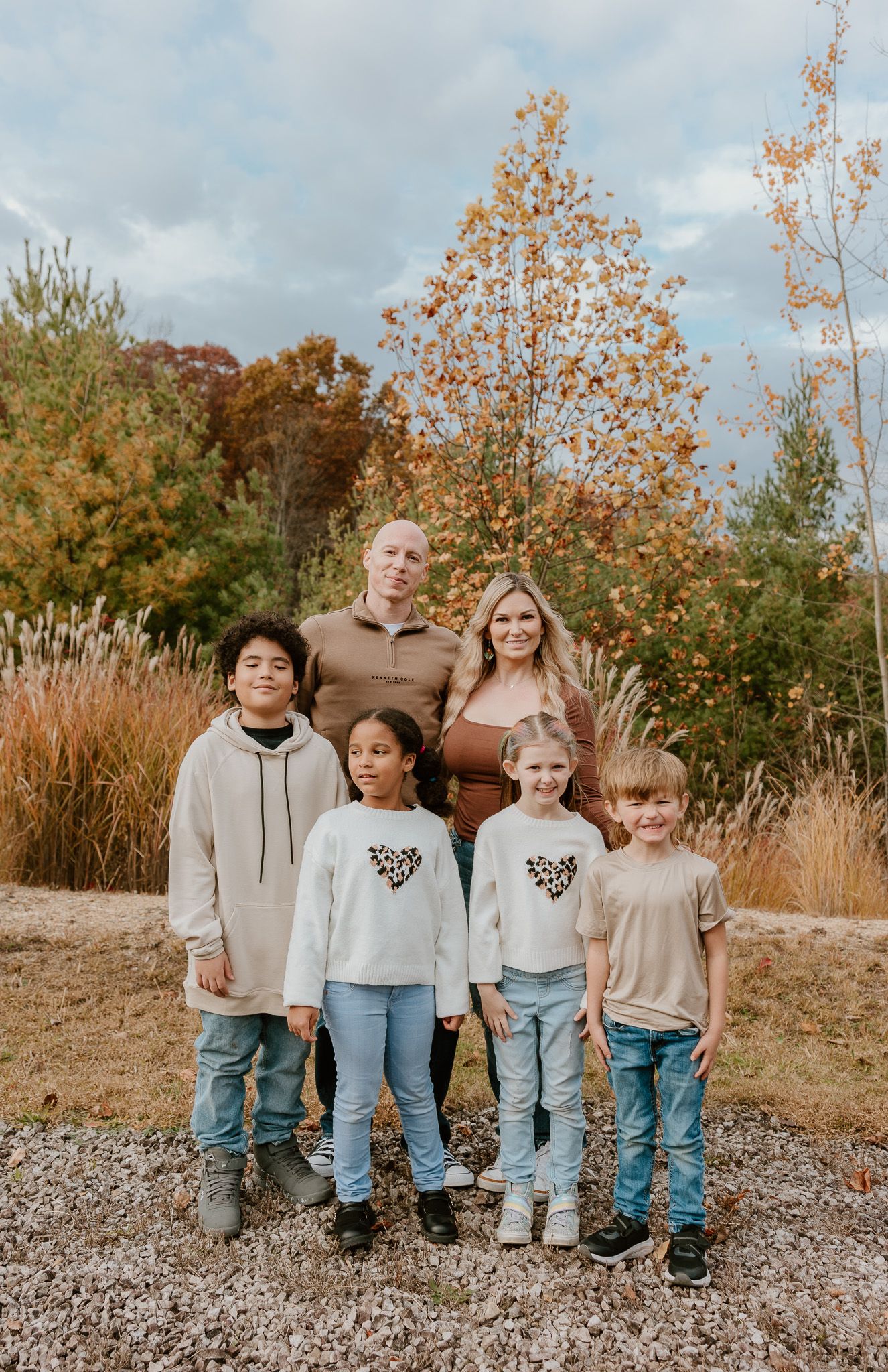 A family poses together outdoors in coordinated casual outfits against a backdrop of fall foliage and tall grasses.