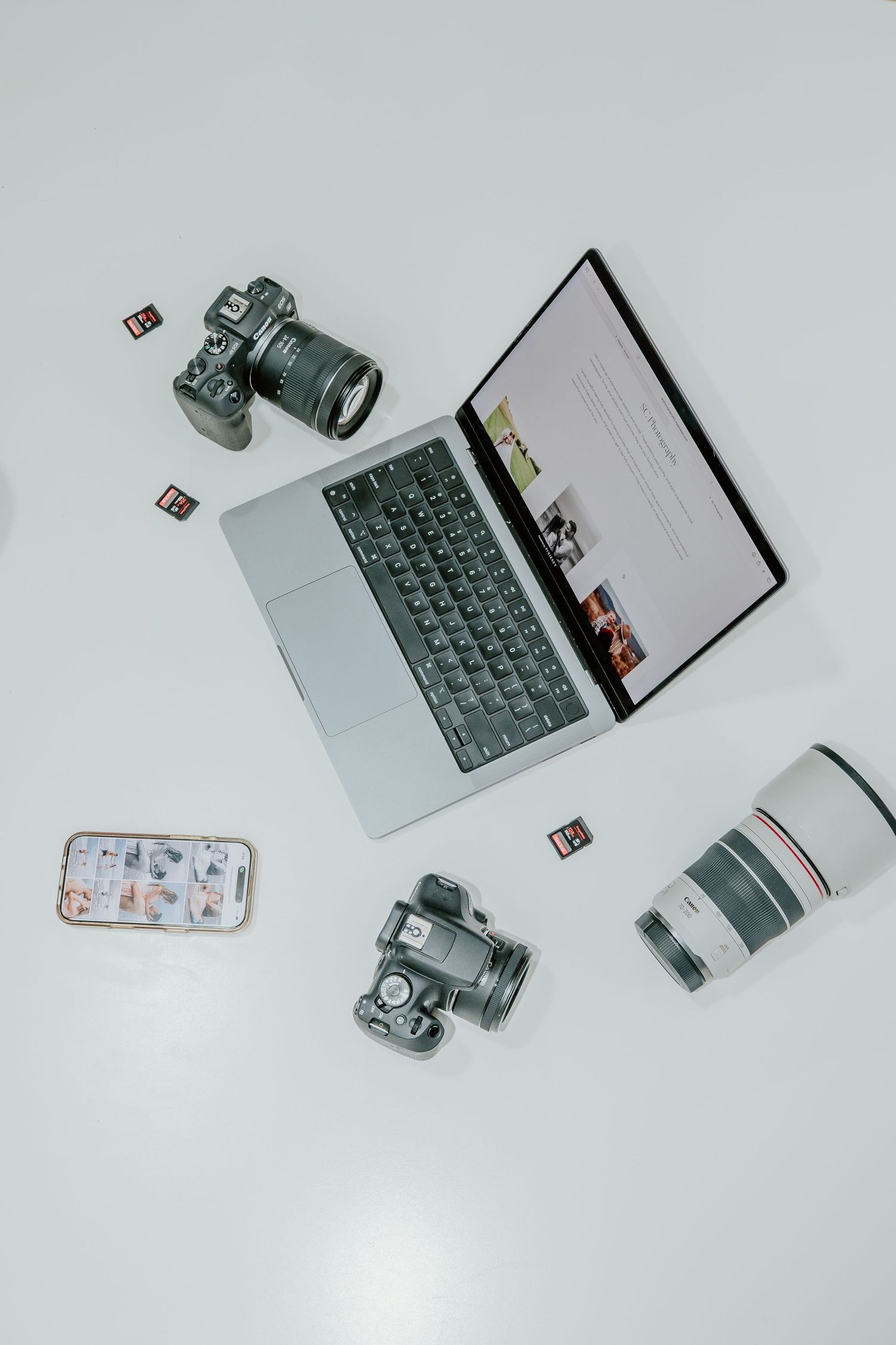 Flatlay of photography equipment including DSLR cameras, laptop, smartphone and camera lenses on white background.