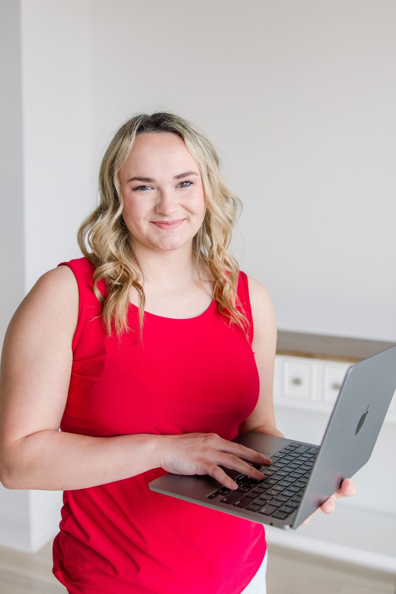 Person in red sleeveless top smiles while working on laptop computer in bright modern space.