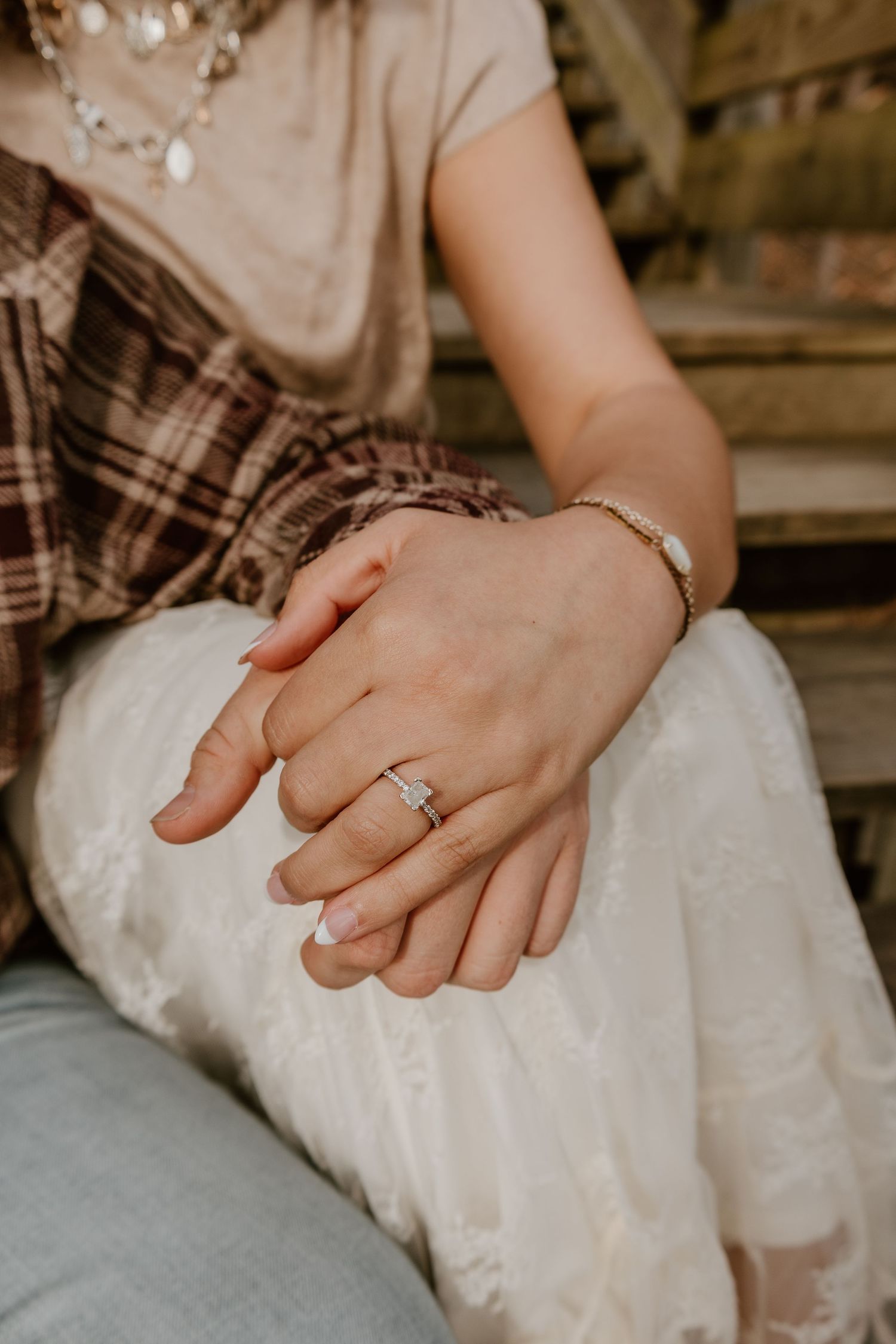 Close-up of hands wearing delicate engagement ring and bracelet resting on white fabric in warm natural lighting.