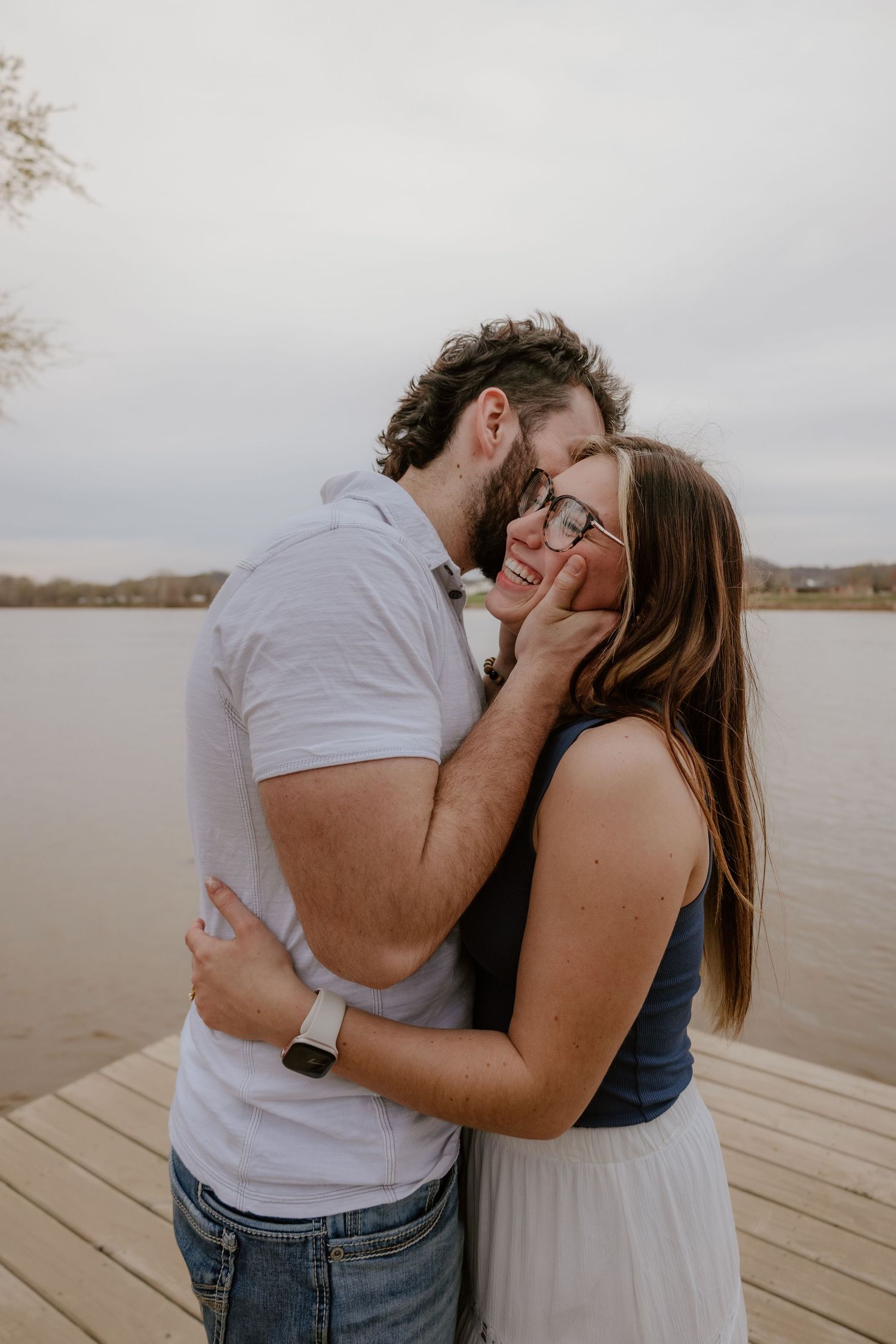 A couple shares a romantic kiss on a wooden dock by a lake on an overcast day.
