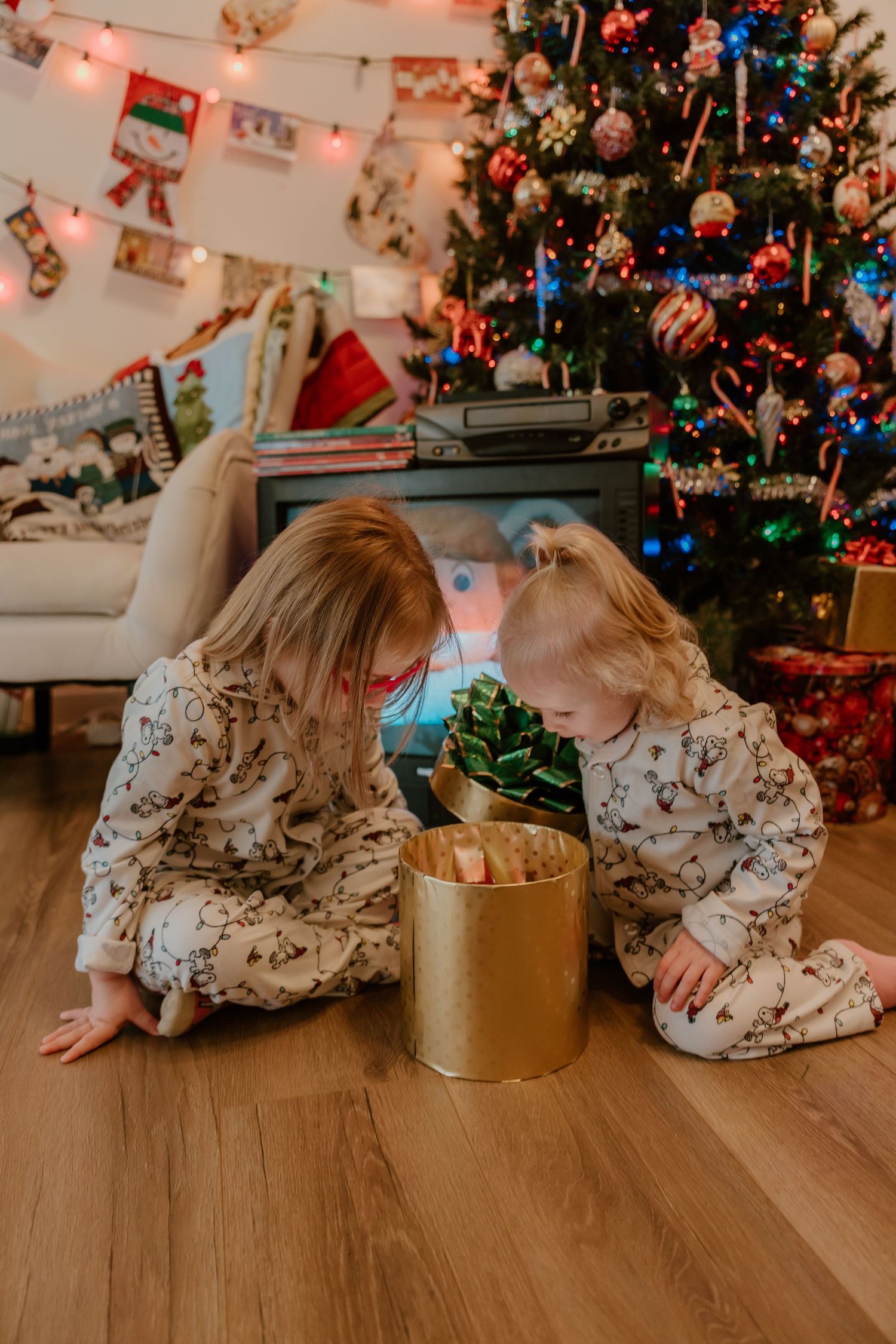 Child in a floral dress crawls across hardwood floor near Christmas tree and gold present box during holiday season.