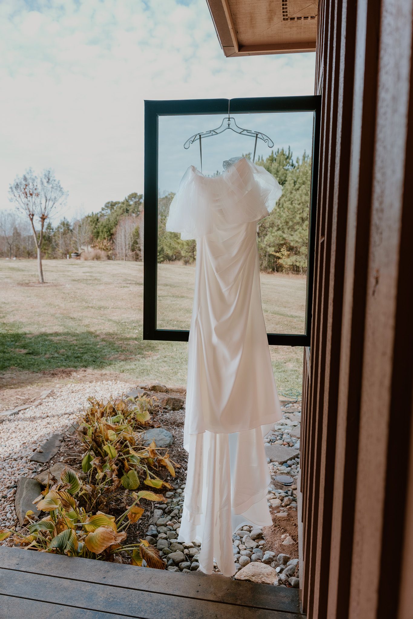 An elegant white wedding dress hangs in a black frame doorway overlooking a natural outdoor landscape.