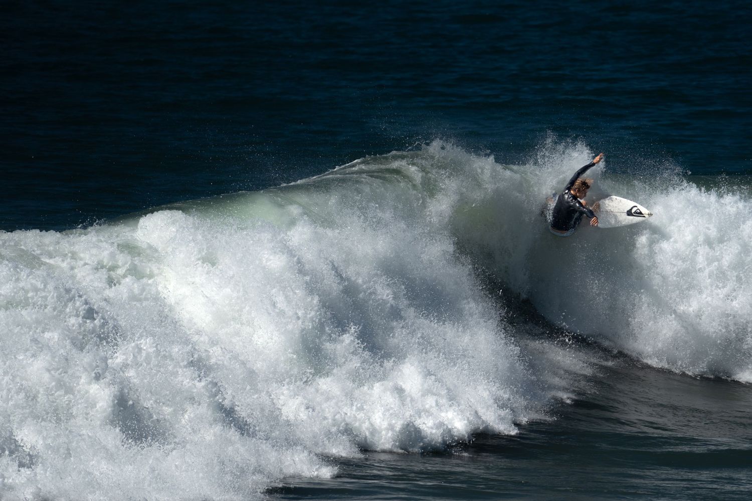 Surfer riding a large crashing ocean wave on a surfboard during a sunny day at the beach.