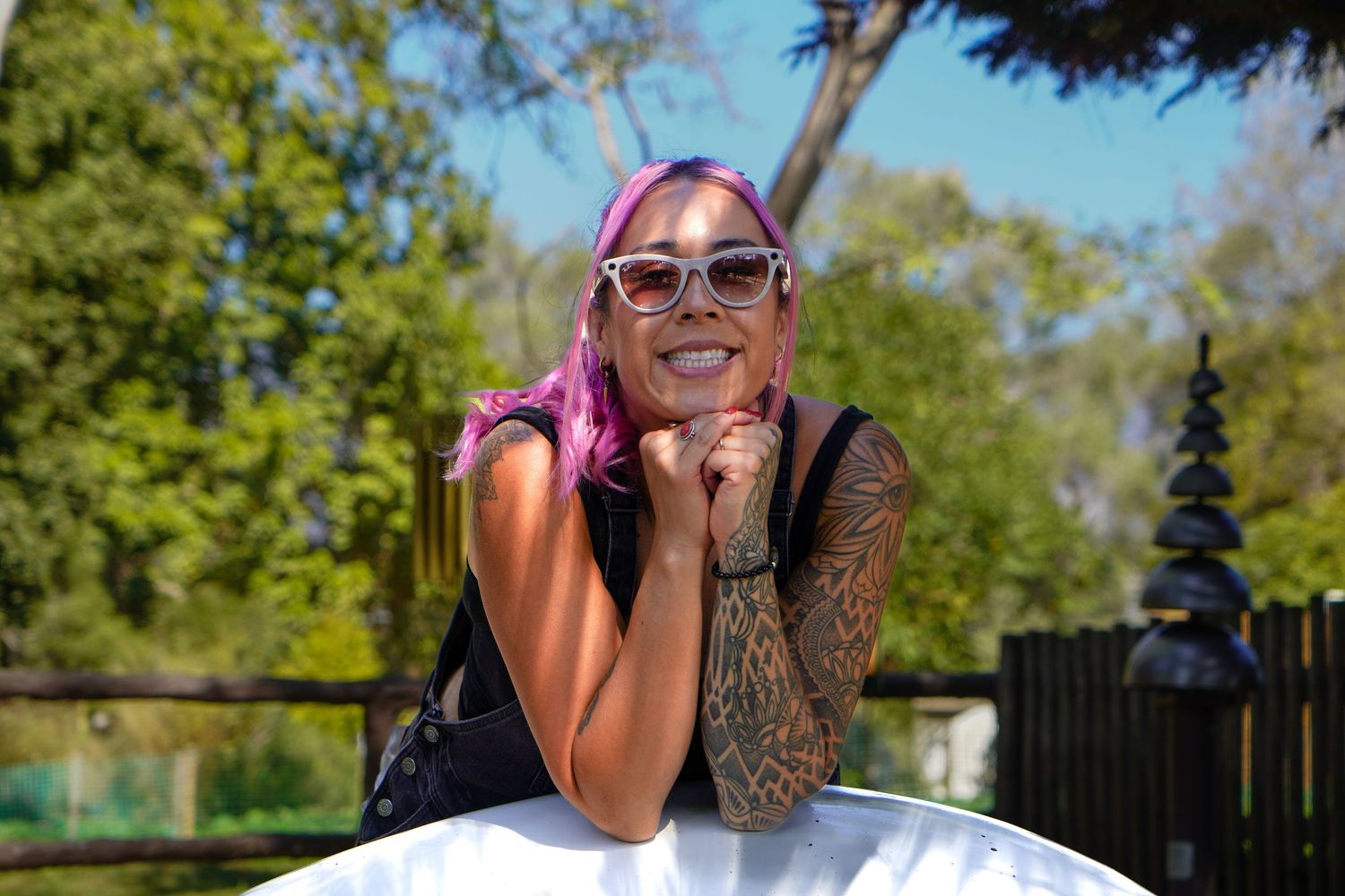 Smiling woman with pink hair, sunglasses, and tattoos posing outdoors on a sunny day near green trees.