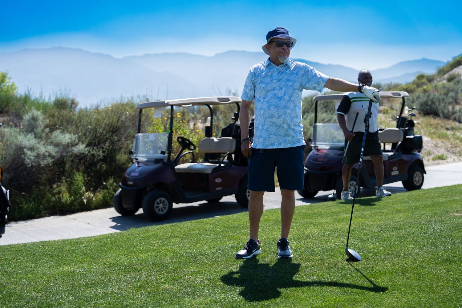 Golfer standing on lush green course holding club, golf carts and mountain landscape in background.