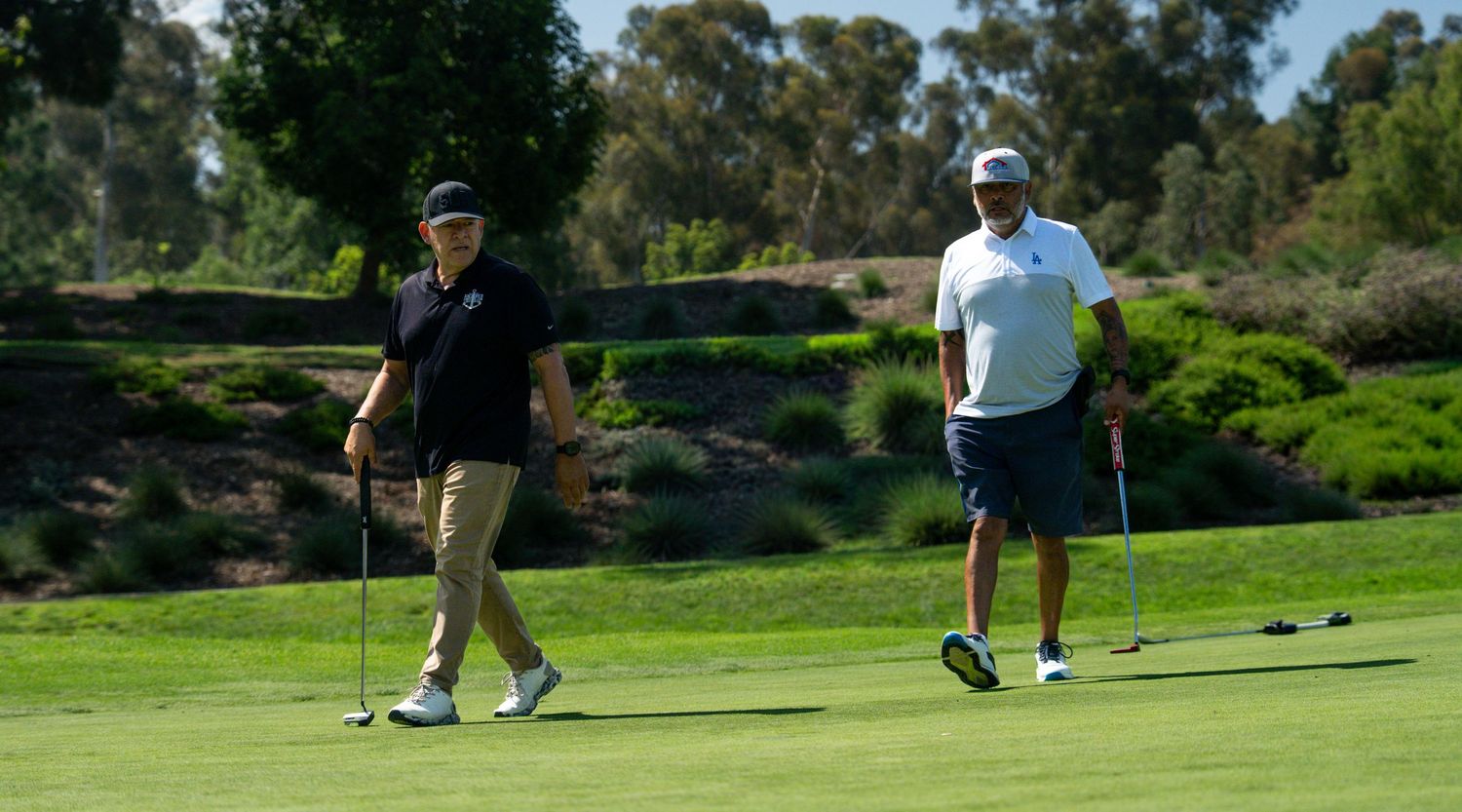 Two golfers walking on a lush green golf course on a sunny day, carrying putters after completing a hole.