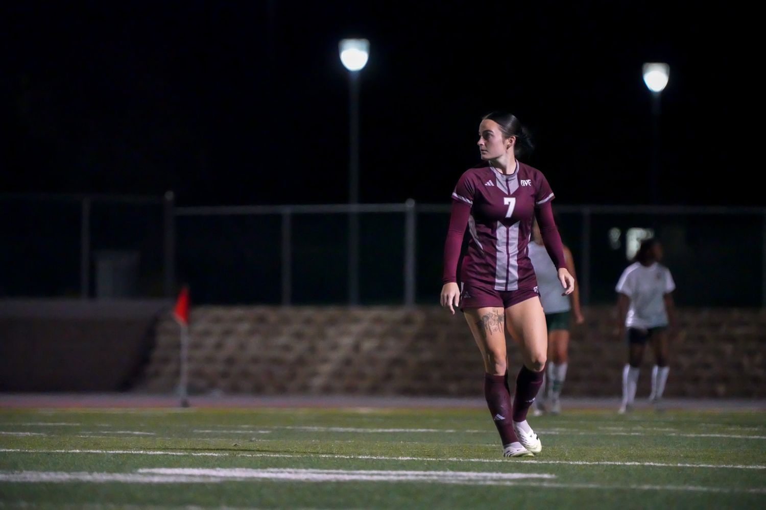 Female soccer player in maroon uniform walking on field during a night game under bright stadium lights.