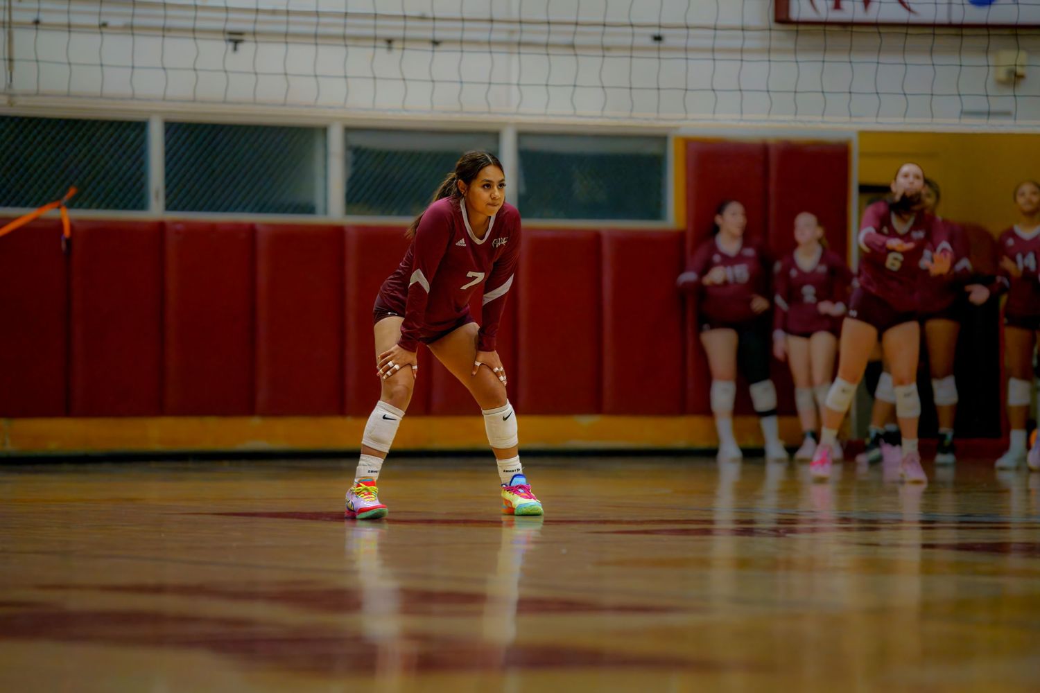 Female volleyball player in maroon uniform crouches in ready position on indoor court during a game.