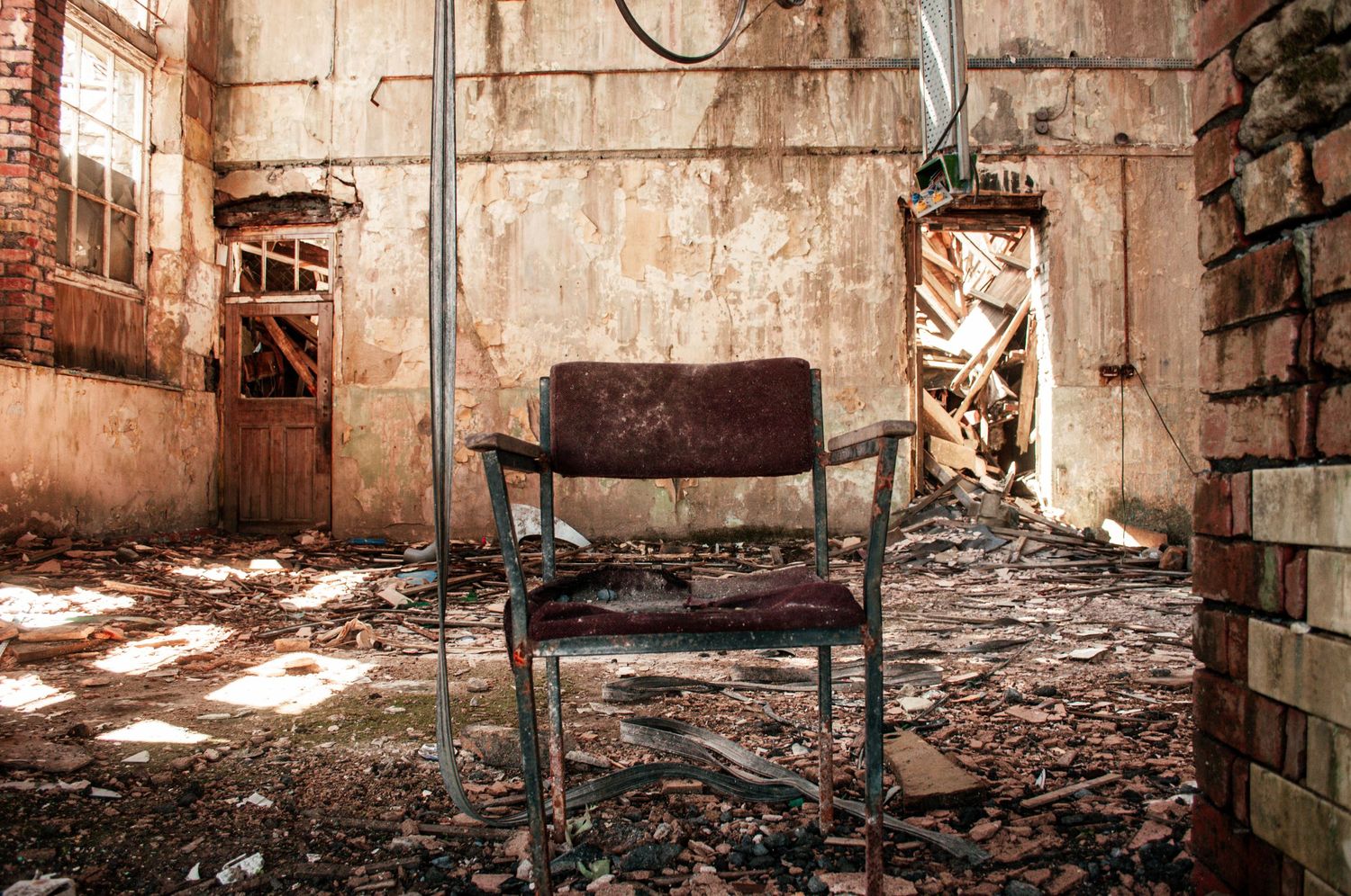 Rusty metal chair in the centre of a decaying industrial room, surrounded by peeling walls, debris, and broken windows. ash olion