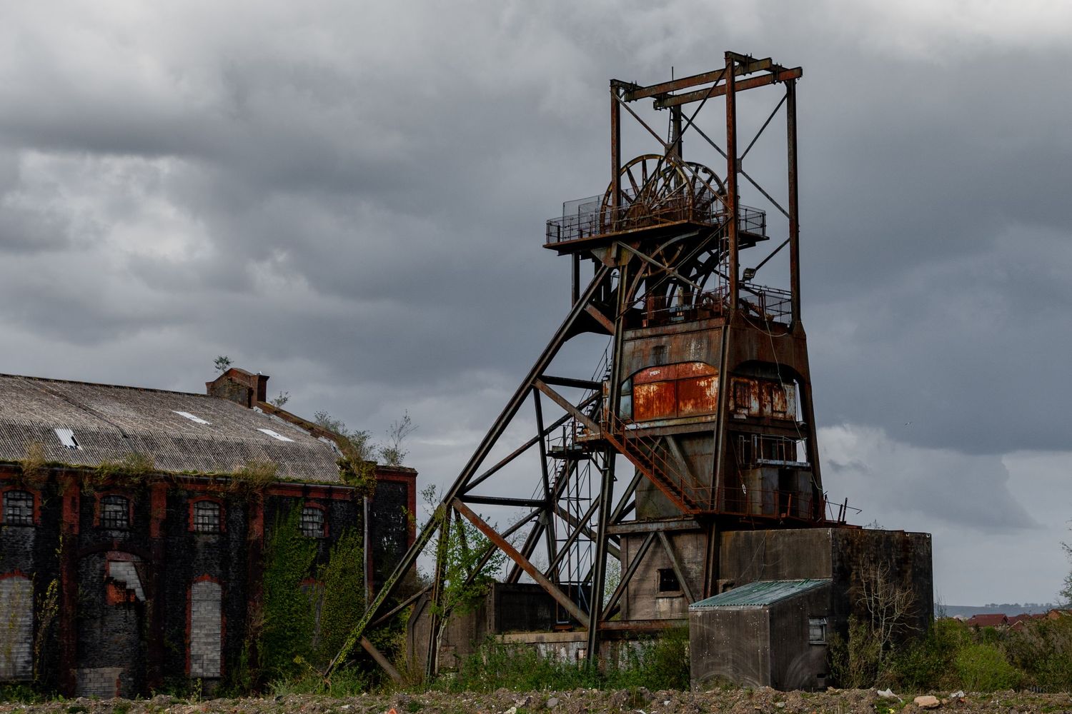 Rusting mining headframe and industrial building beneath a heavy overcast sky. penallta colliery. ash olion photography. derelict wales