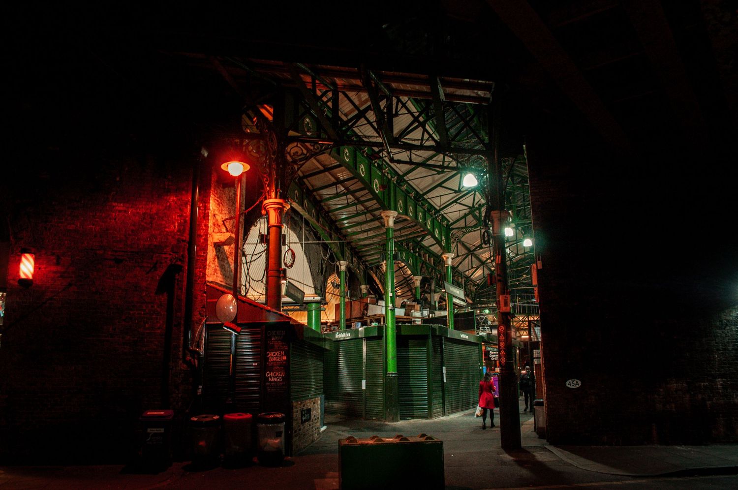 Night-time view inside a covered market hall, with warm orange and green lights illuminating the metal beams and stalls london