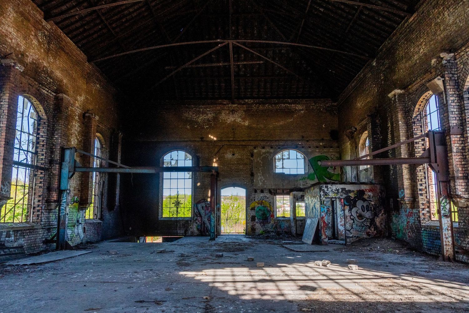 Wide view of a derelict brick hall with tall arched windows, scattered debris, and strong sunlight streaming across the floor.  ash olion