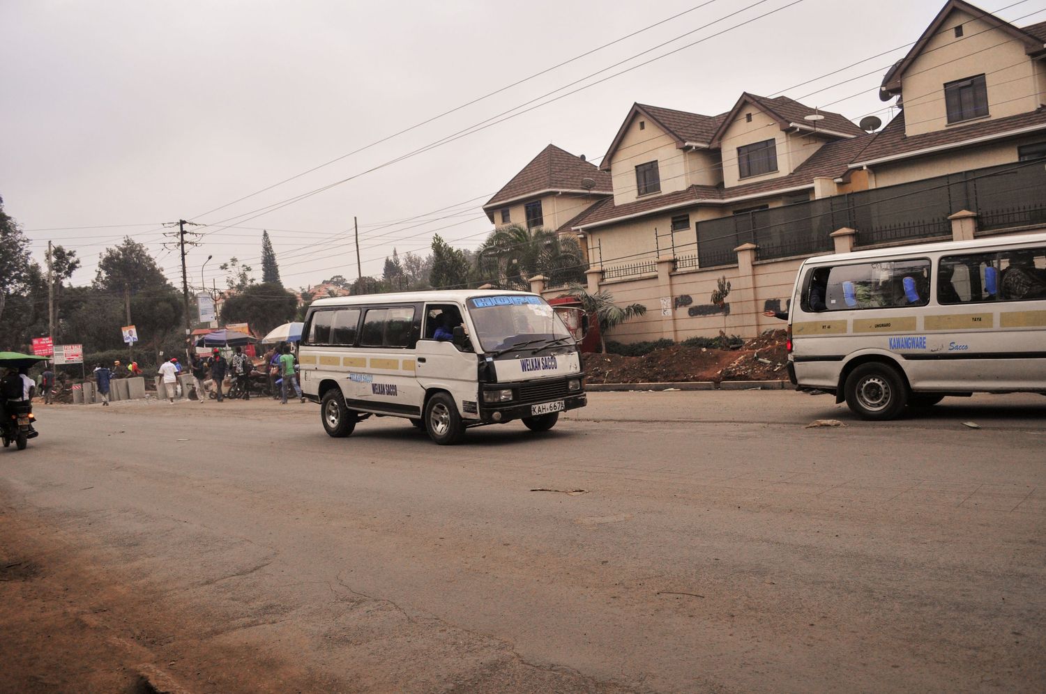 Street scene with two white minibuses driving past a row of large houses under an overcast sky. Kenya, Africa