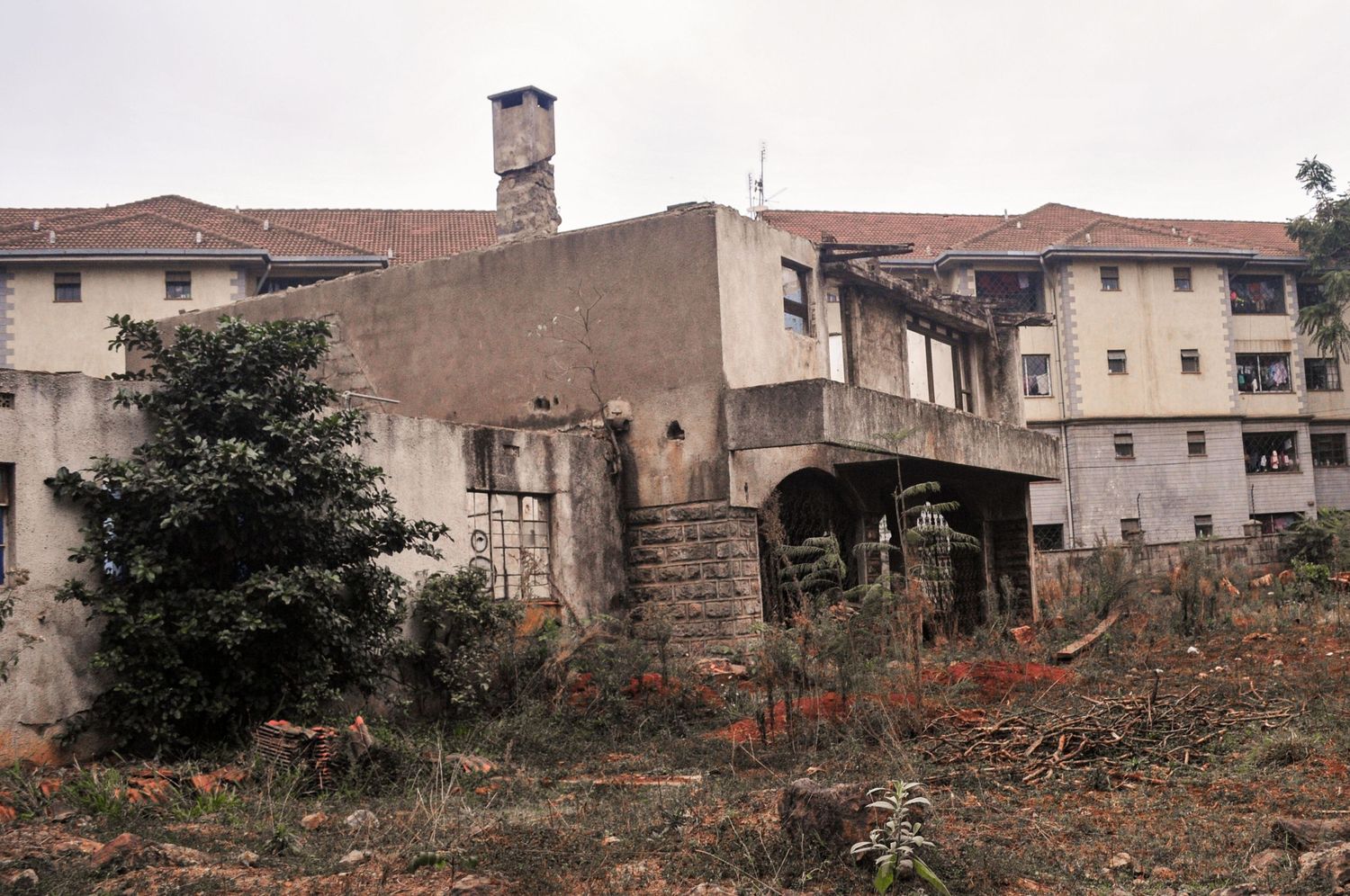 Derelict multi-storey building with peeling paint and broken windows, surrounded by an overgrown, neglected yard. Kenya, Africa
