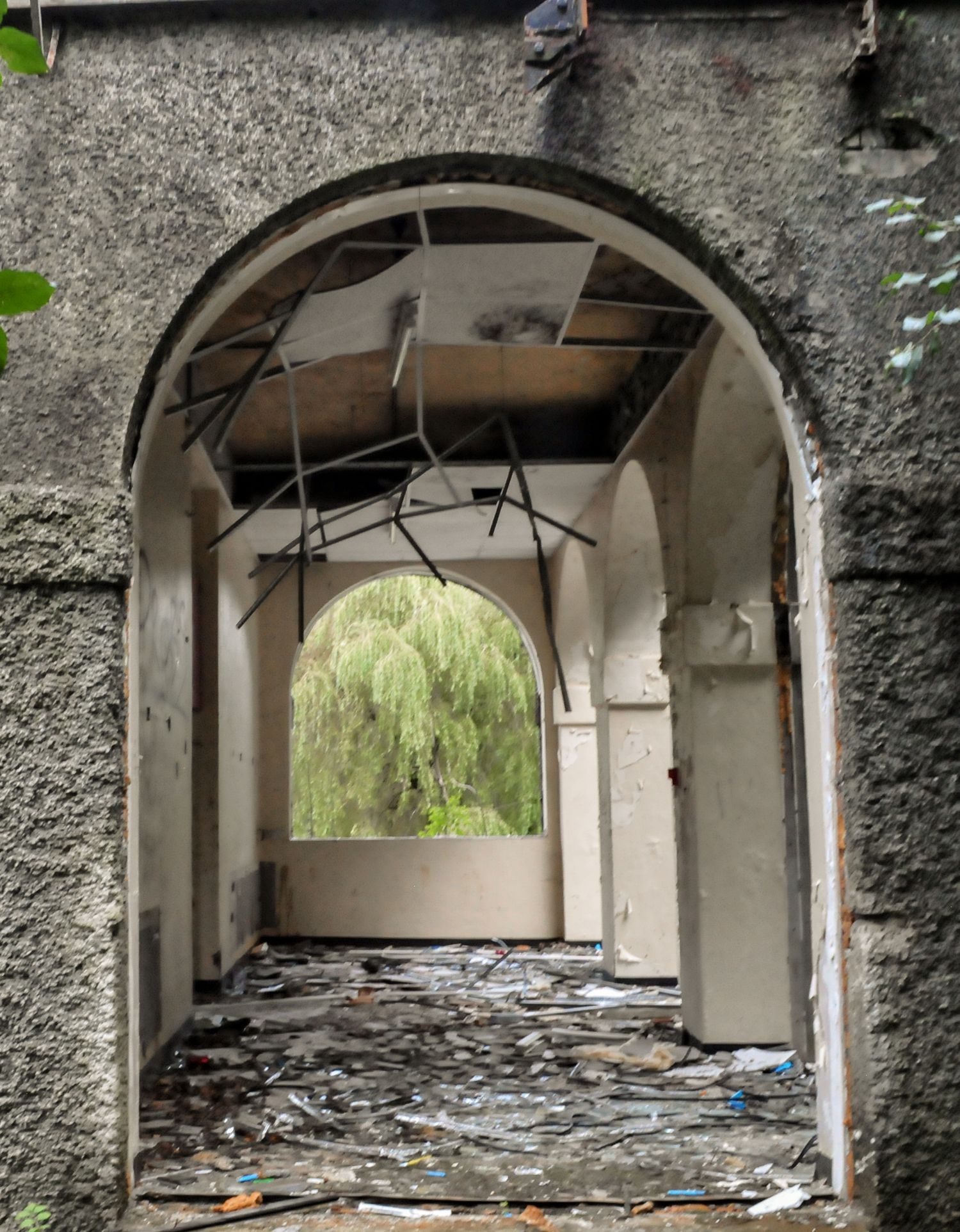 Long abandoned corridor framed by repeating stone arches, with debris covering the floor and bright greenery visible at the far end.