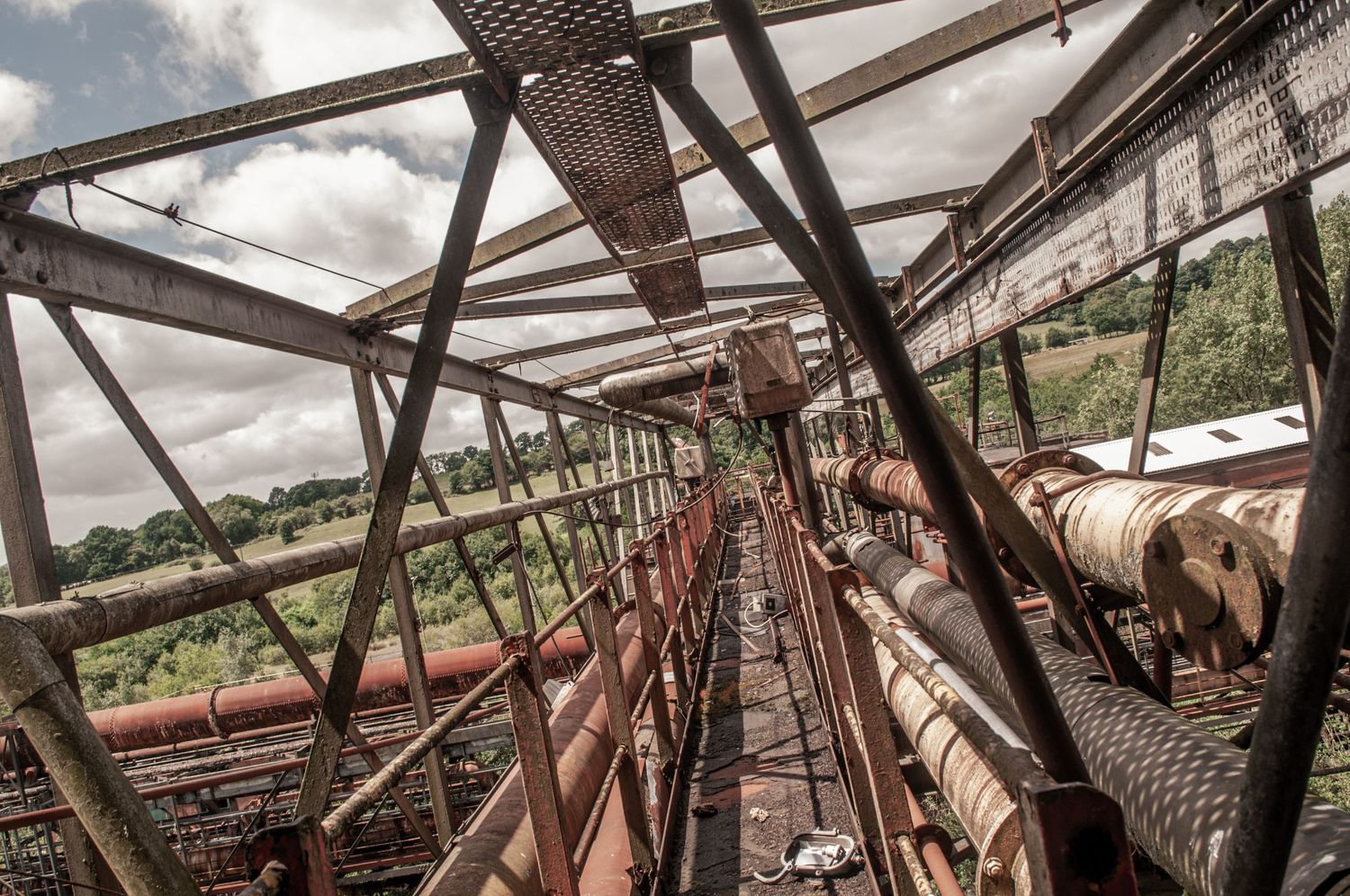 ash olion. View down a rusted metal walkway enclosed by steel beams, overlooking long industrial pipes. cwm coke works beddau