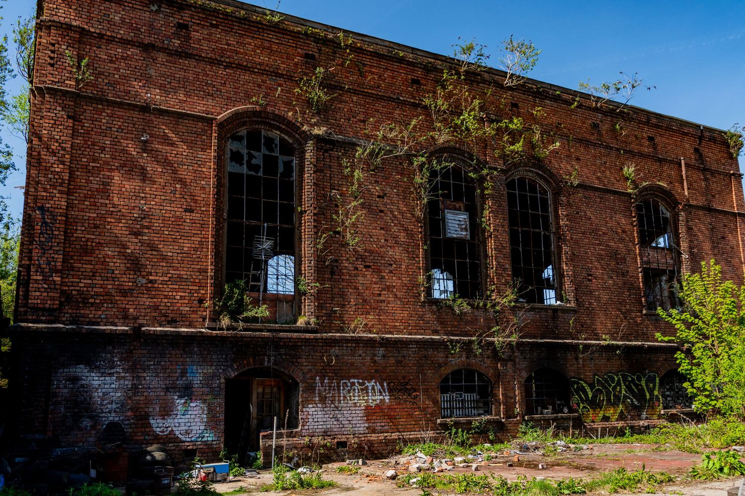 Exterior of a derelict red-brick industrial building with tall arched windows, overgrown foliage, and signs of long-term decay. wales