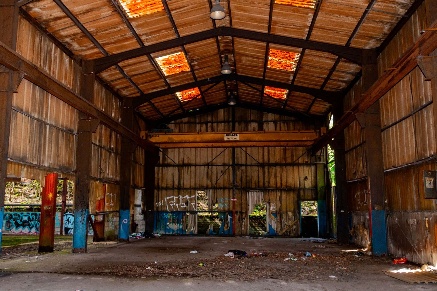 Interior of a long, rusted corrugated-metal warehouse with broken panels, scattered debris, and graffiti on the far wall. derelict wales