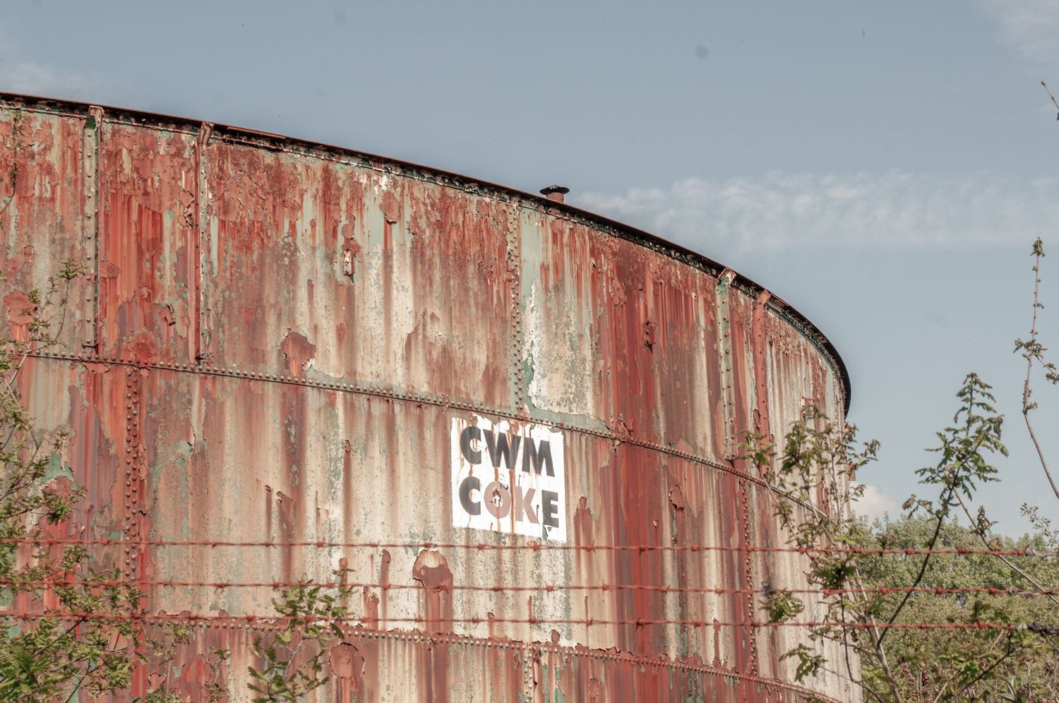 Large rusted metal storage tank marked with faded lettering, partially hidden by surrounding greenery. cwm coke works beddau wales.