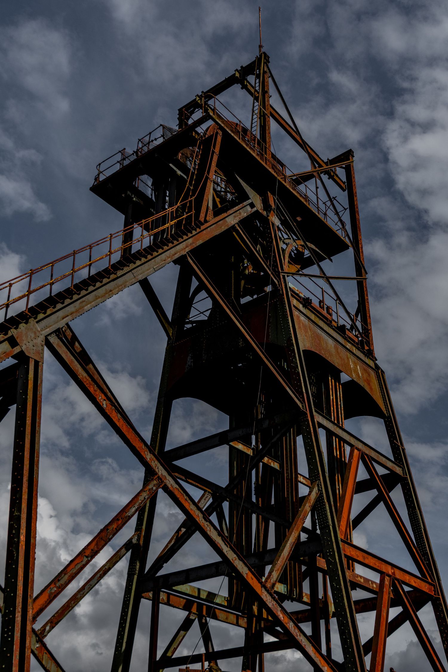Rusted steel mining headstock rising against a cloudy sky, with angular beams and elevated walkways. penallta colliery ash olion wales