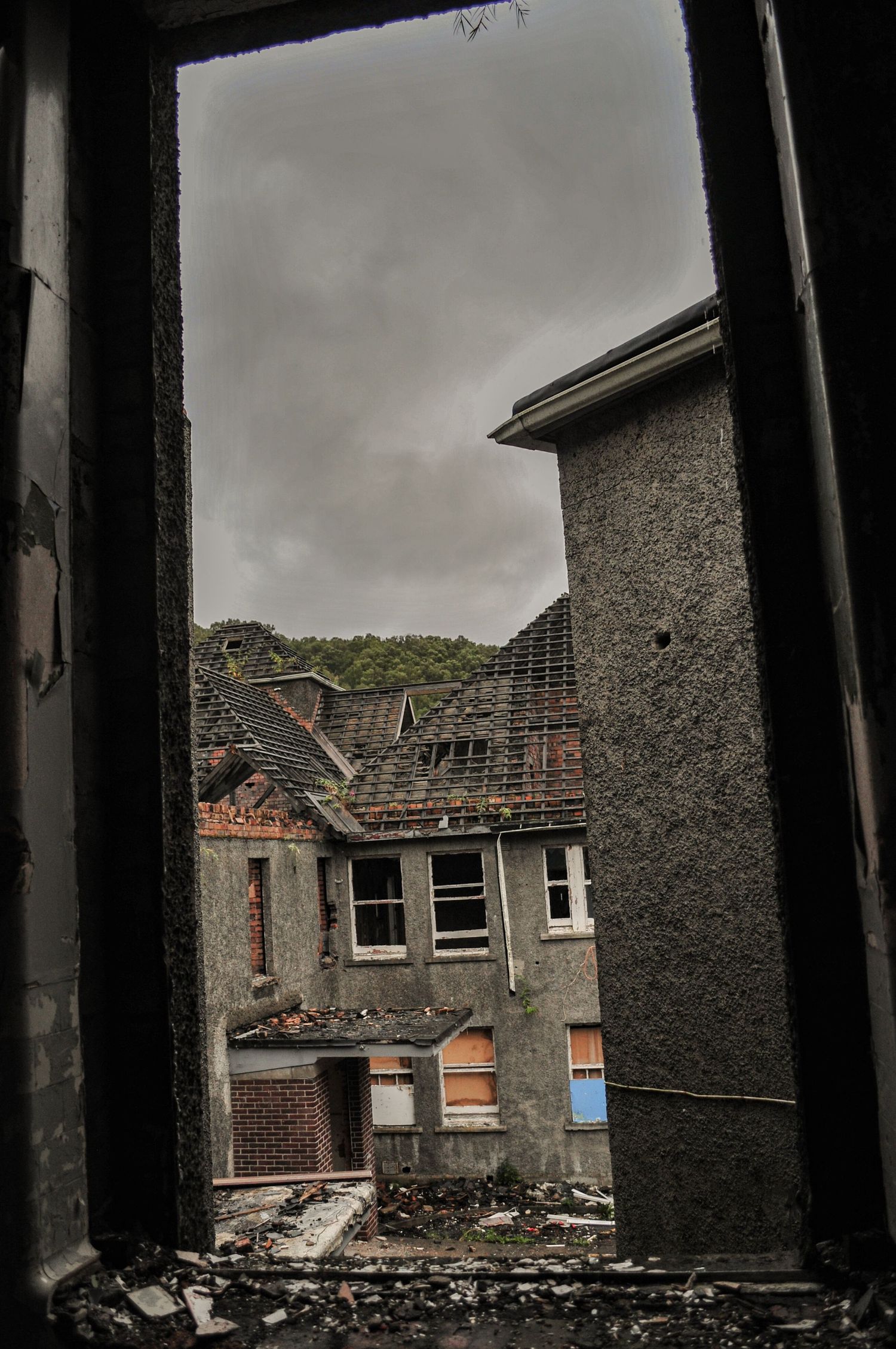 View through a darkened window frame looking out onto a courtyard of derelict stone buildings under an overcast sky. mountain ash hospital