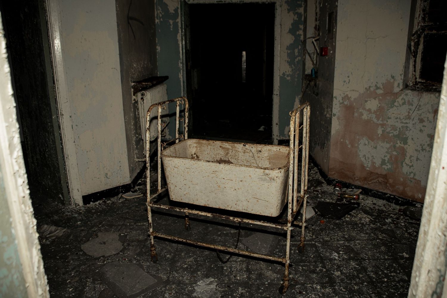 Old metal cot positioned near a black doorway in a decayed room, surrounded by peeling walls and broken flooring. amman valley maternity