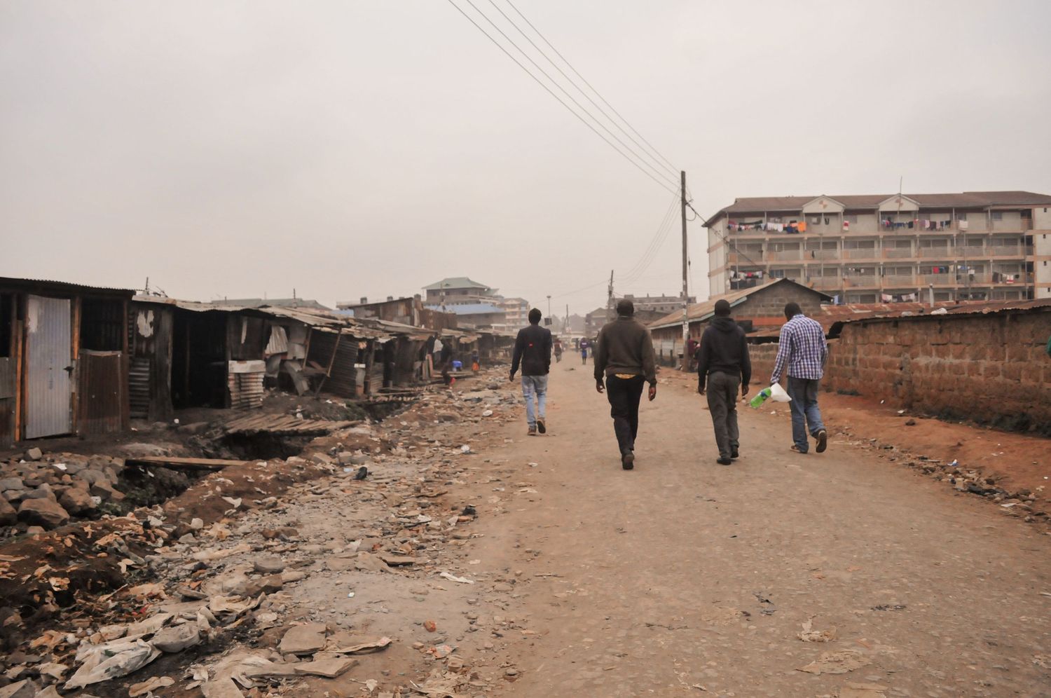 Group of people walking down a dusty roadside lined with buildings, with larger apartment blocks visible in the distance. ash olion. kenya