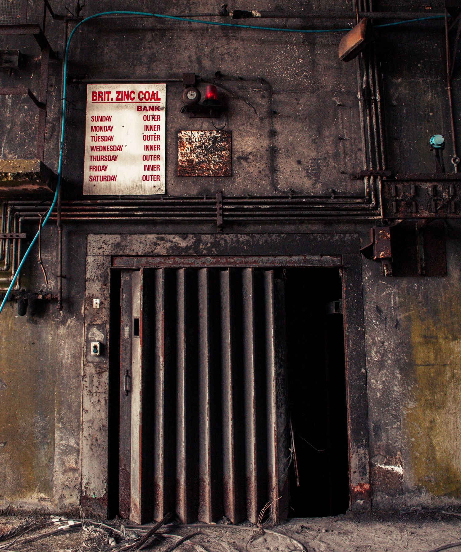 ash olion Dark industrial entrance with vertical metal bars, surrounded by peeling walls, exposed pipes, and a faded instruction sign above