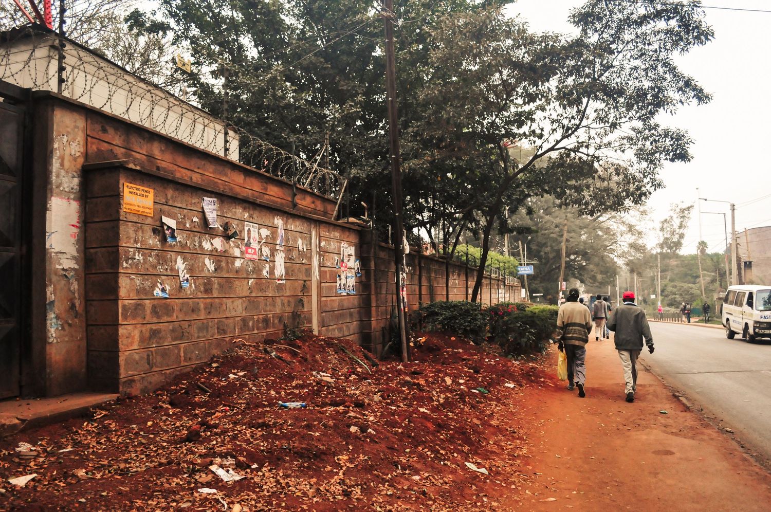 Street view of people walking along a red dirt roadside beside a weathered brick wall plastered with old posters. ash olion Kenya, Africa
