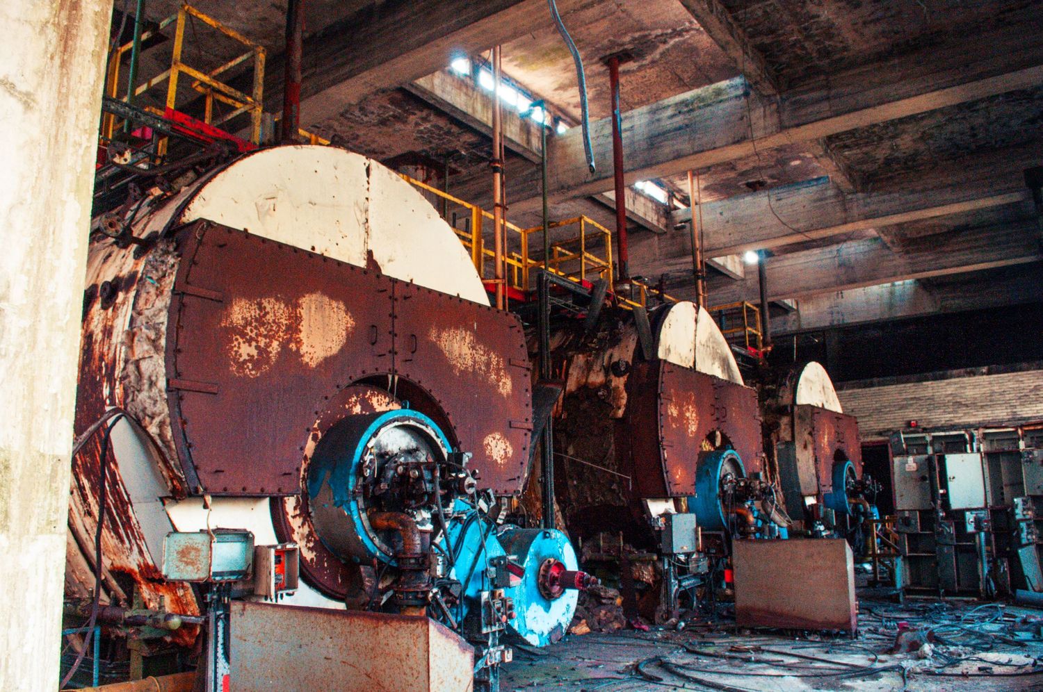 Row of massive rusted industrial boilers inside a derelict factory hall, scattered with rubble and peeling paint. cwm coke works beddau
