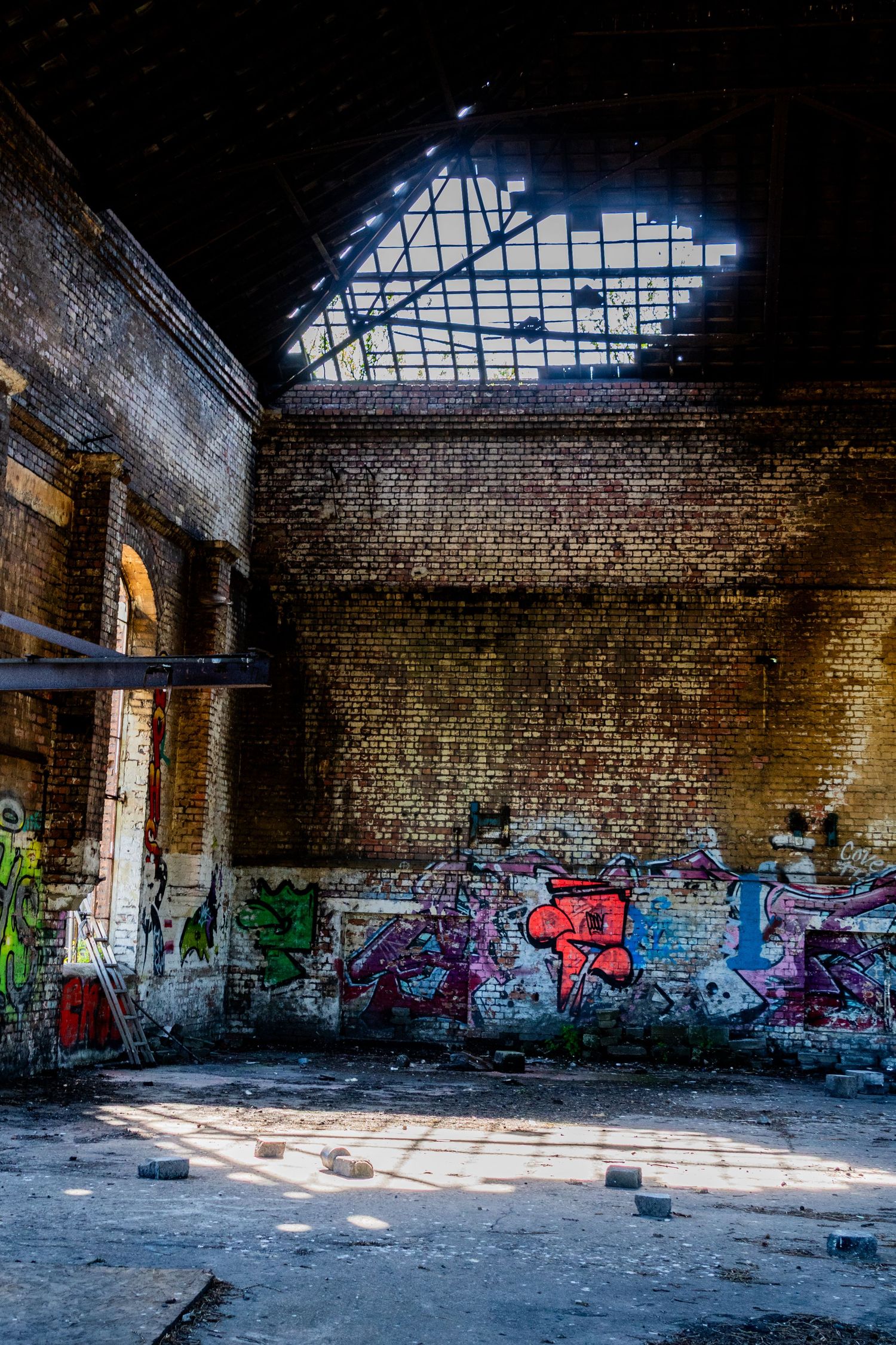 Large abandoned industrial hall with a broken roof letting in sunlight, illuminating graffiti-covered walls and scattered debris. ash olion