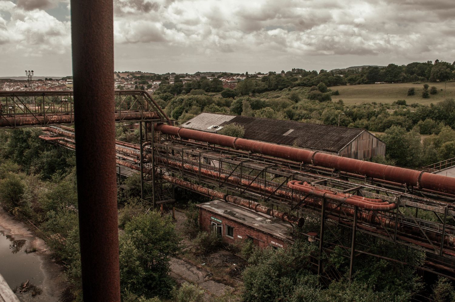 ash olion Elevated view of long rusted industrial pipes stretching across an overgrown landscape beneath a cloudy sky. cwm coke works beddau