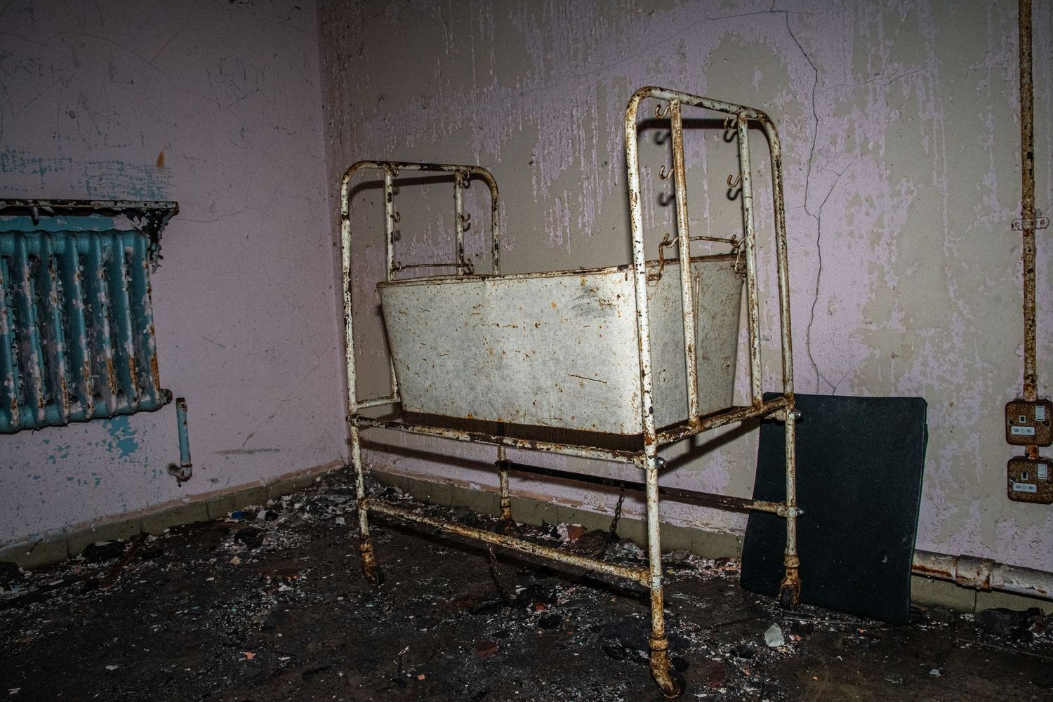 Old metal hospital crib in a peeling, derelict room with debris scattered on the floor. amman valley maternity hospital ash olion photo