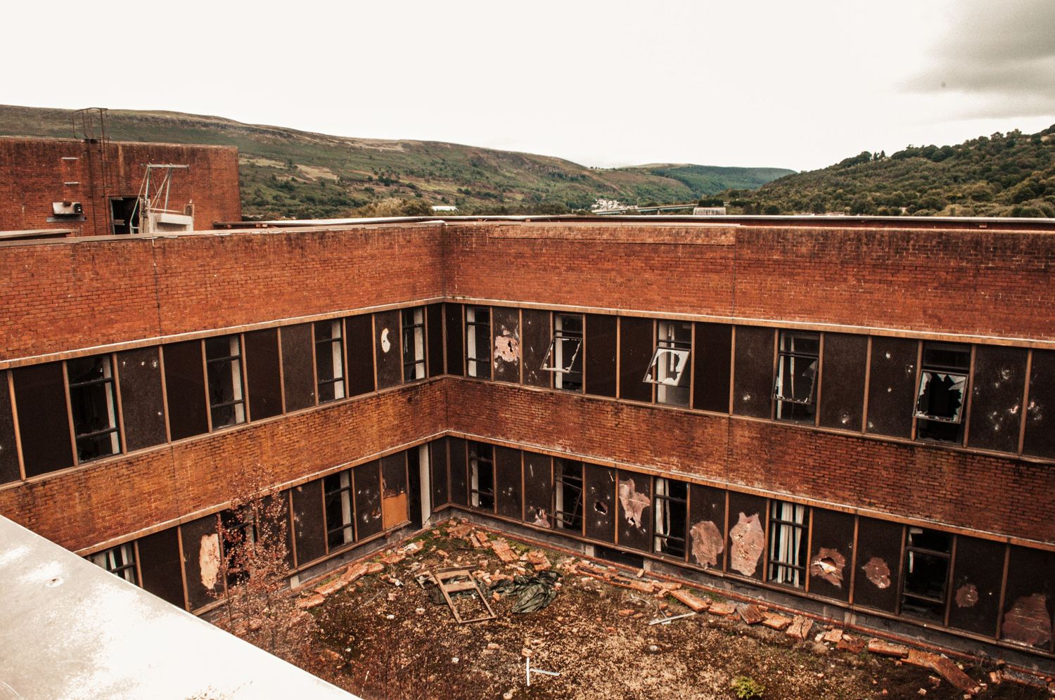 Abandoned square building with broken windows surrounding an overgrown central courtyard, set against rolling hills. hoover factory merthyr