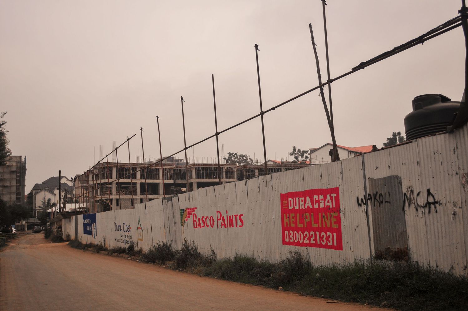 Long roadside construction wall covered with painted adverts and signs, running alongside a dirt street .Kenya, Africa
