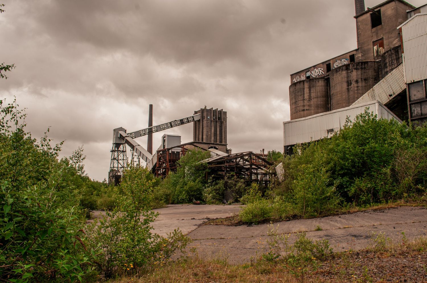 Crumbling industrial buildings surrounded by overgrown vegetation beneath a dark, cloud-filled sky. cwm coke works beddau. derelict abandon