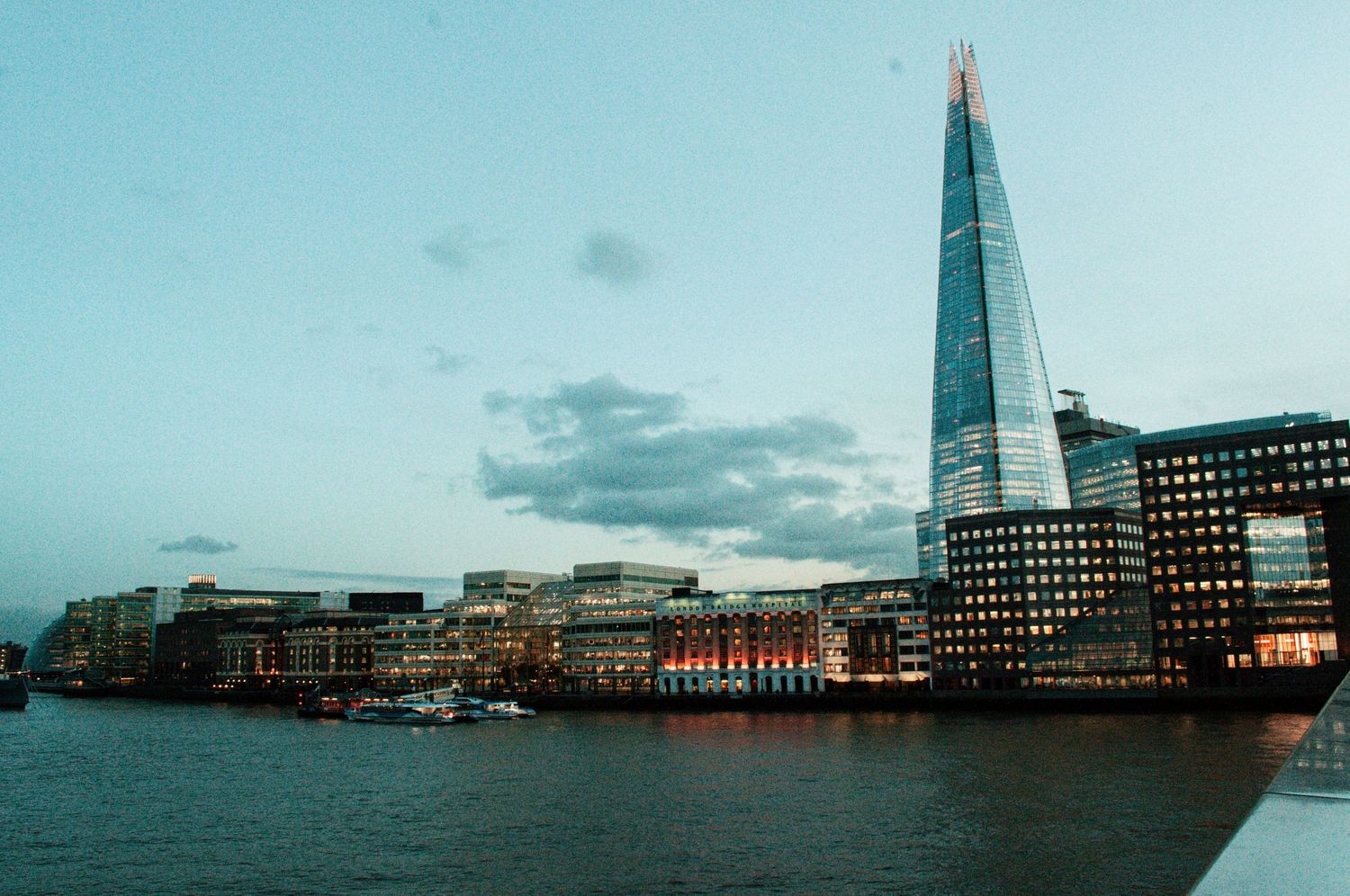 Wide view of the London skyline featuring the Shard rising above the Thames beneath a pale blue, cloud-strewn sky