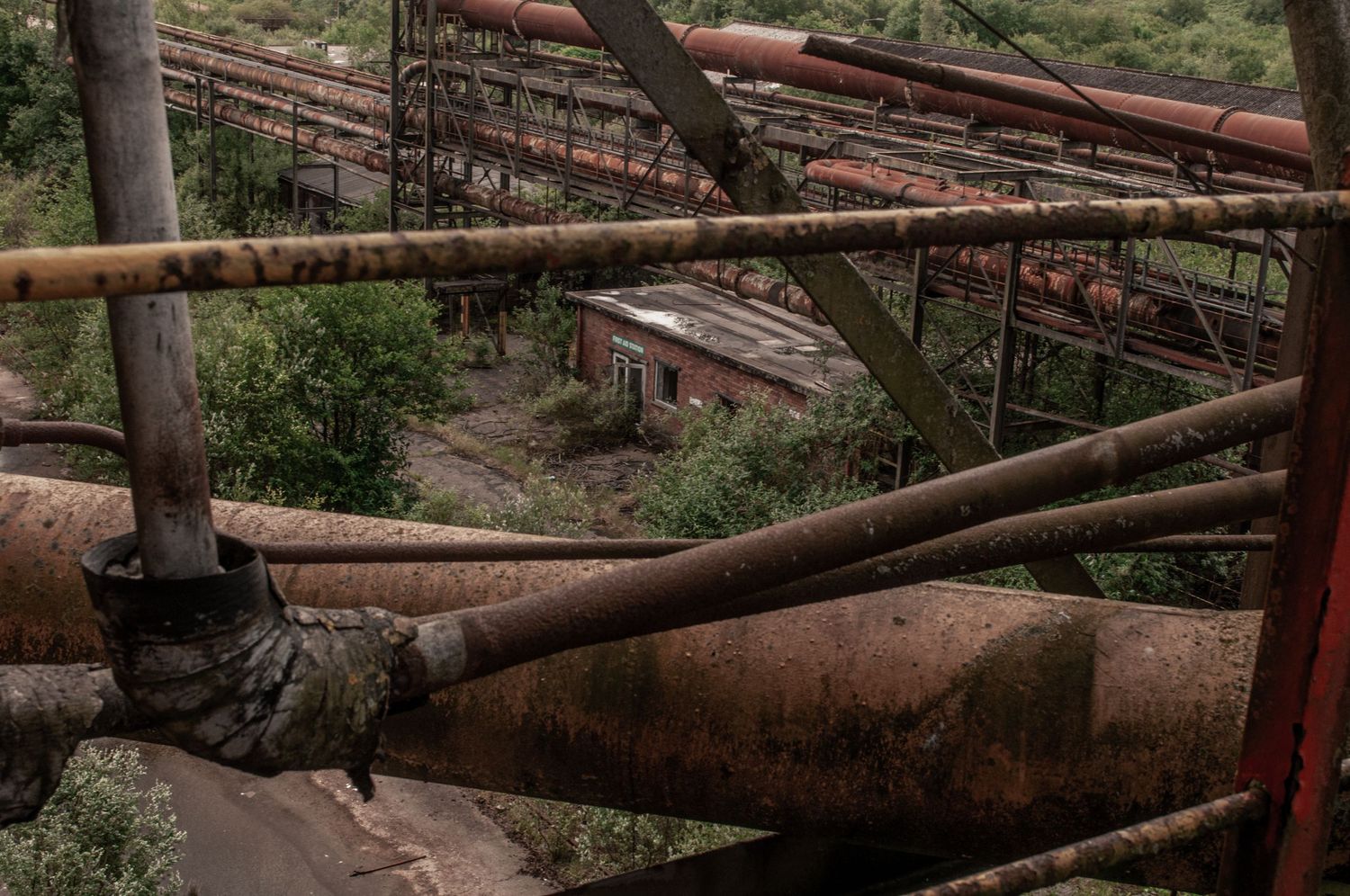 ash olion.Network of rusted metal pipes and railings overlooking an overgrown industrial site reclaimed by vegetation. cwm coke works