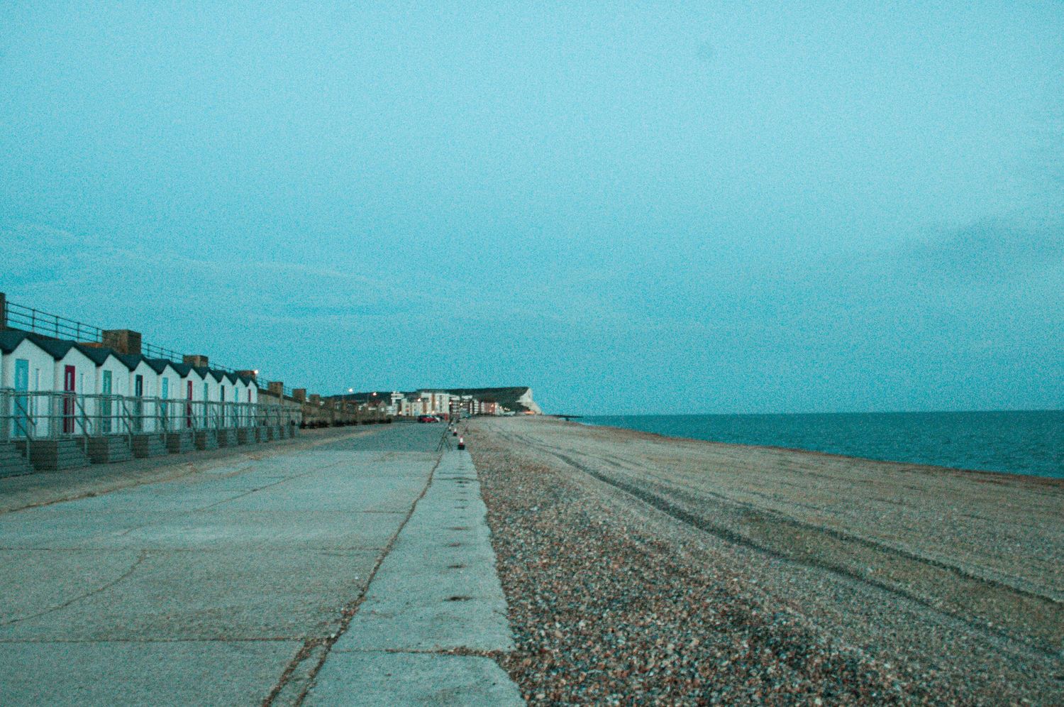 Wide coastal view of a quiet promenade beside a pebble beach, with a row of white beach huts pale blue sky. Newhaven