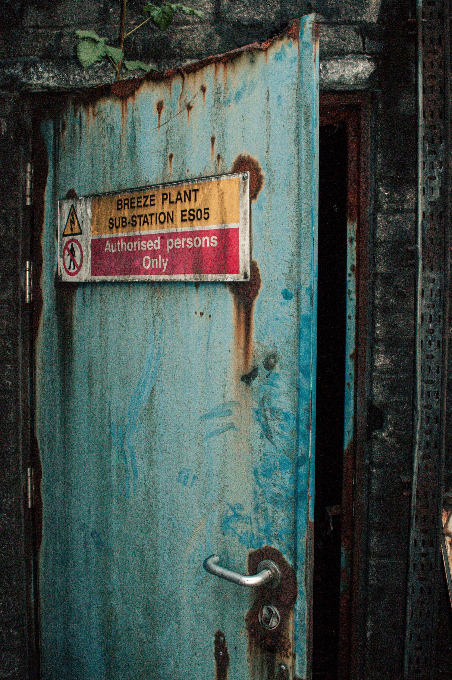 Weathered blue metal door marked with a faded warning sign, its surface rusted and partially open into a dark industrial interior ash olion