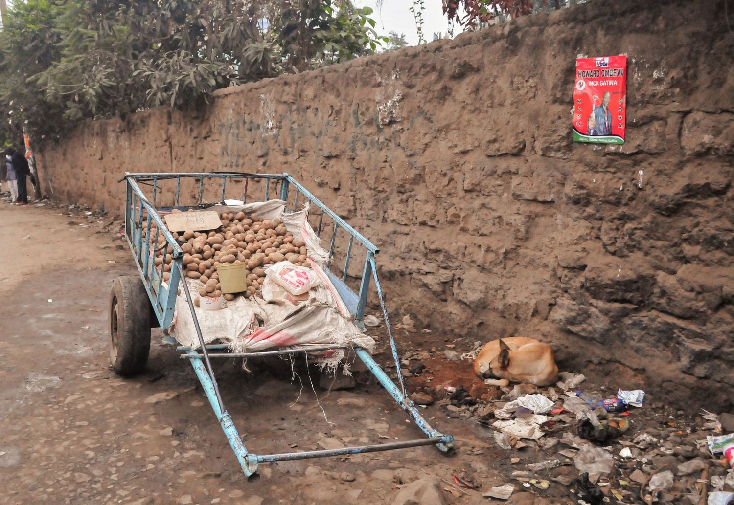 A metal handcart filled with rubble sits guarded by a dog on a dirt roadside next to a stone retaining wall. ash olion. kenya, africa