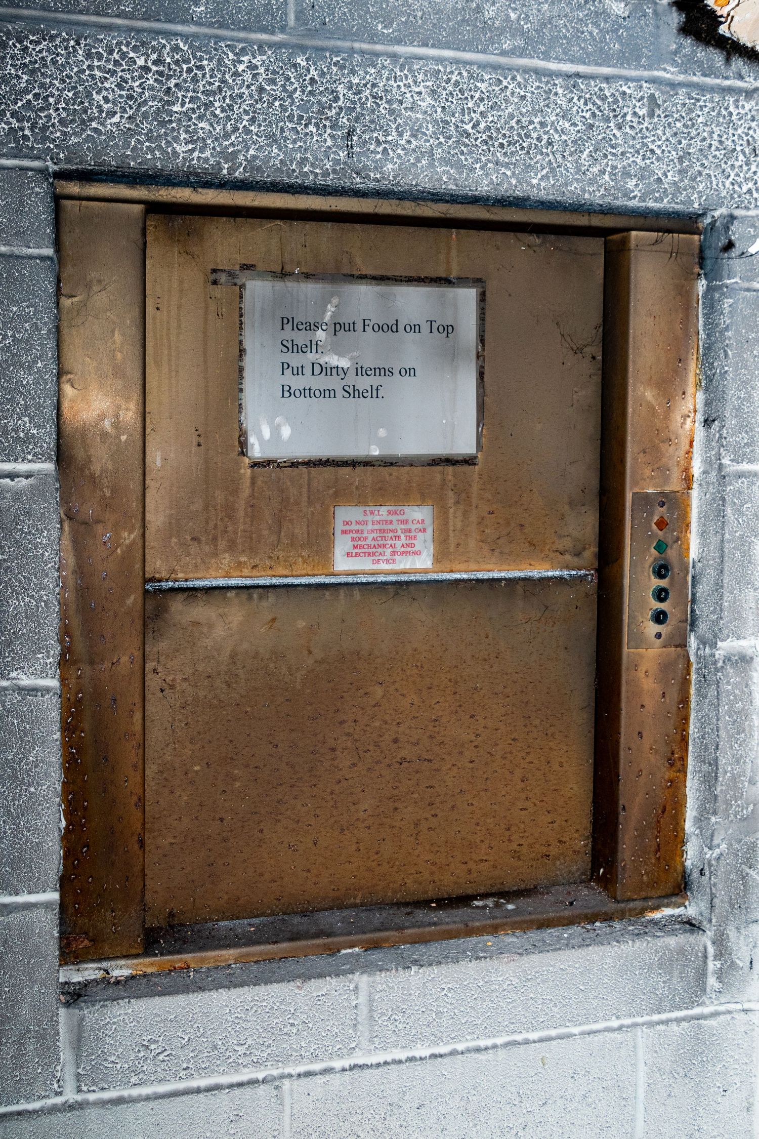 Old metal service hatch set into a stone wall, with a faded sign and tarnished metal fixtures.  spin bowling cardiff. urbex abandoned