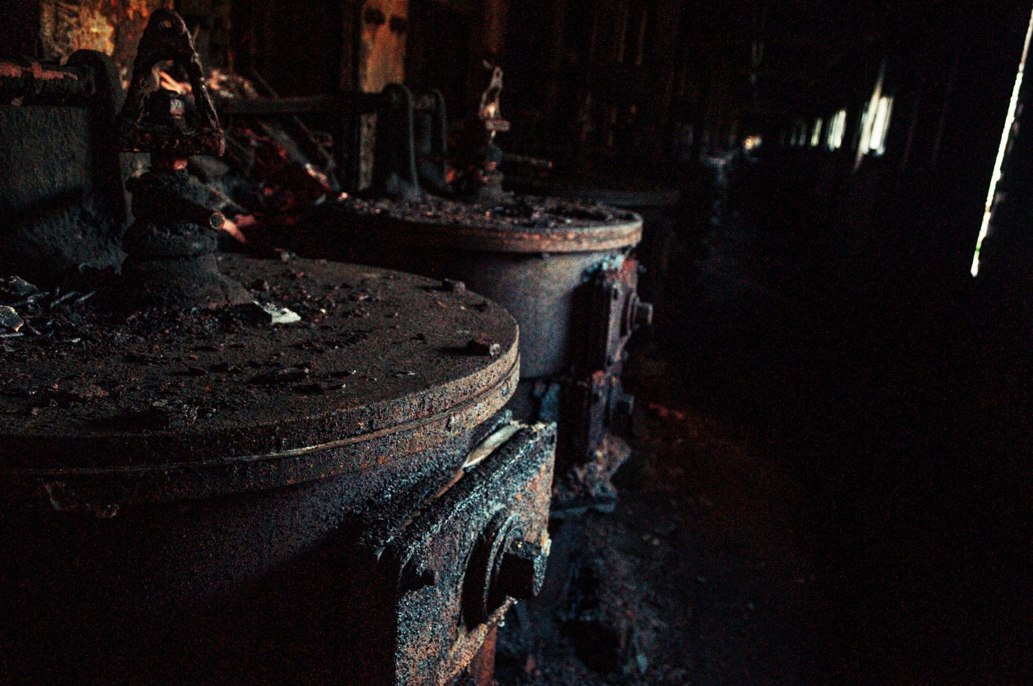 Dimly lit industrial room with large circular machinery covered in rust and debris, fading into darkness.  cwm coke works beddau. ash olion