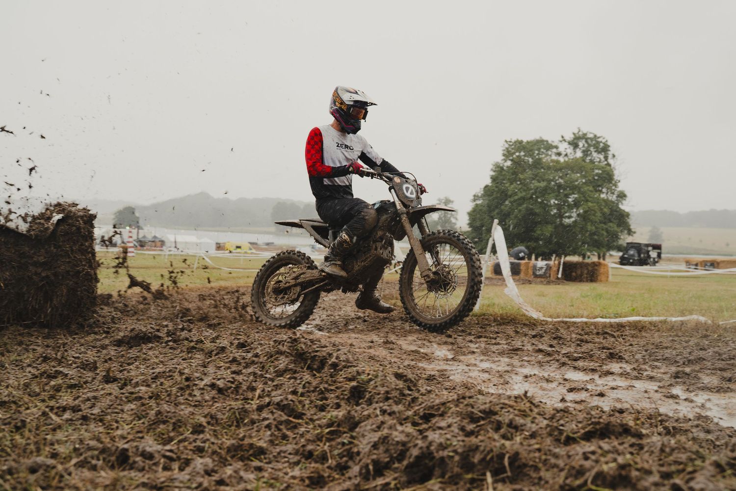 Dirt bike riders traversing muddy terrain in a rural landscape.