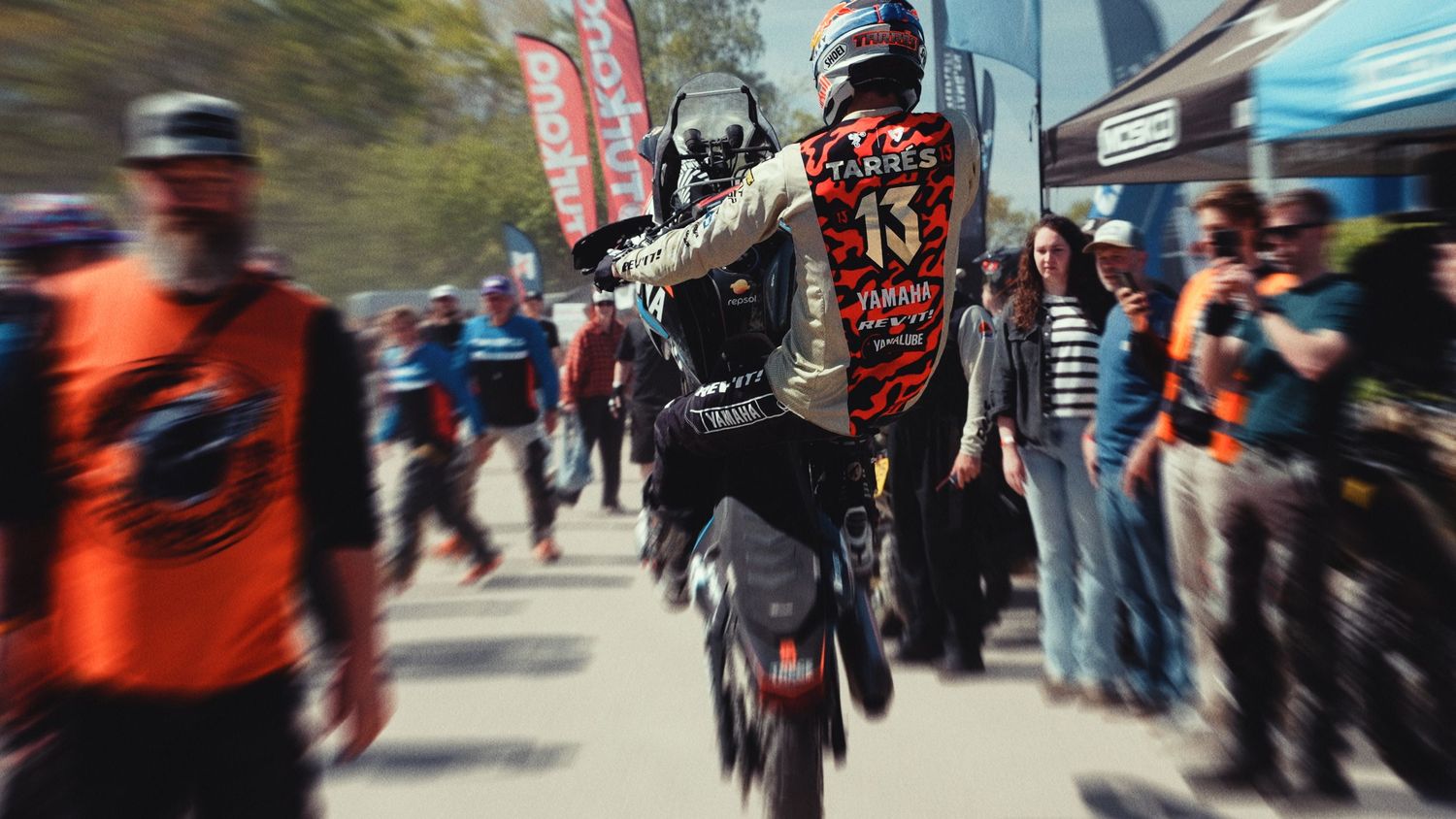 A motocross rider performs a wheelie on a dirt bike through a crowded outdoor event with spectators watching.