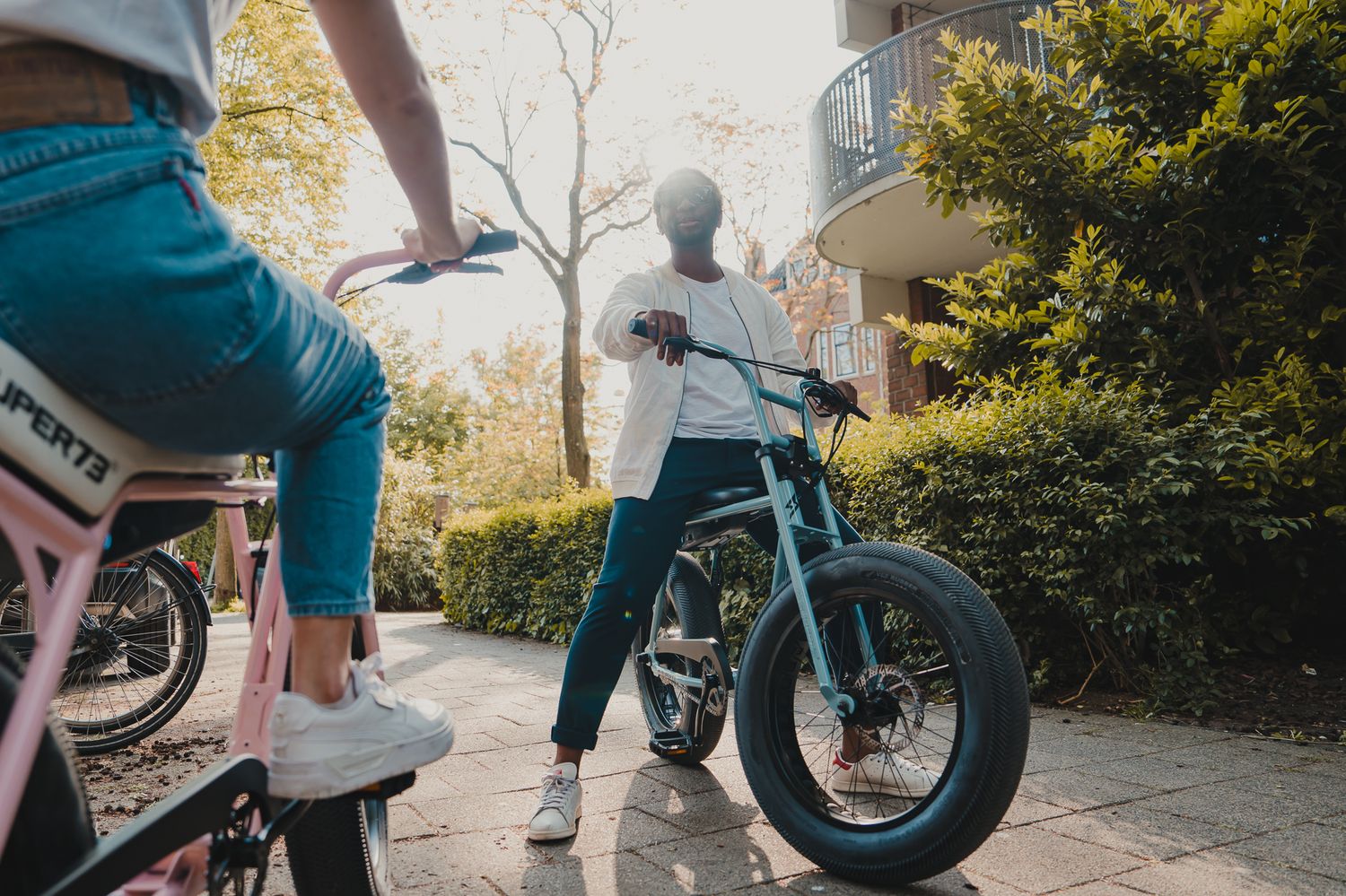 Two people riding stylish electric bikes through a sunny outdoor area with trees in the background.
