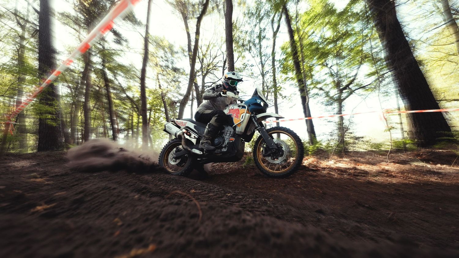 Motocross rider racing through a forest dirt track, kicking up mud with trees and orange course markers in background.